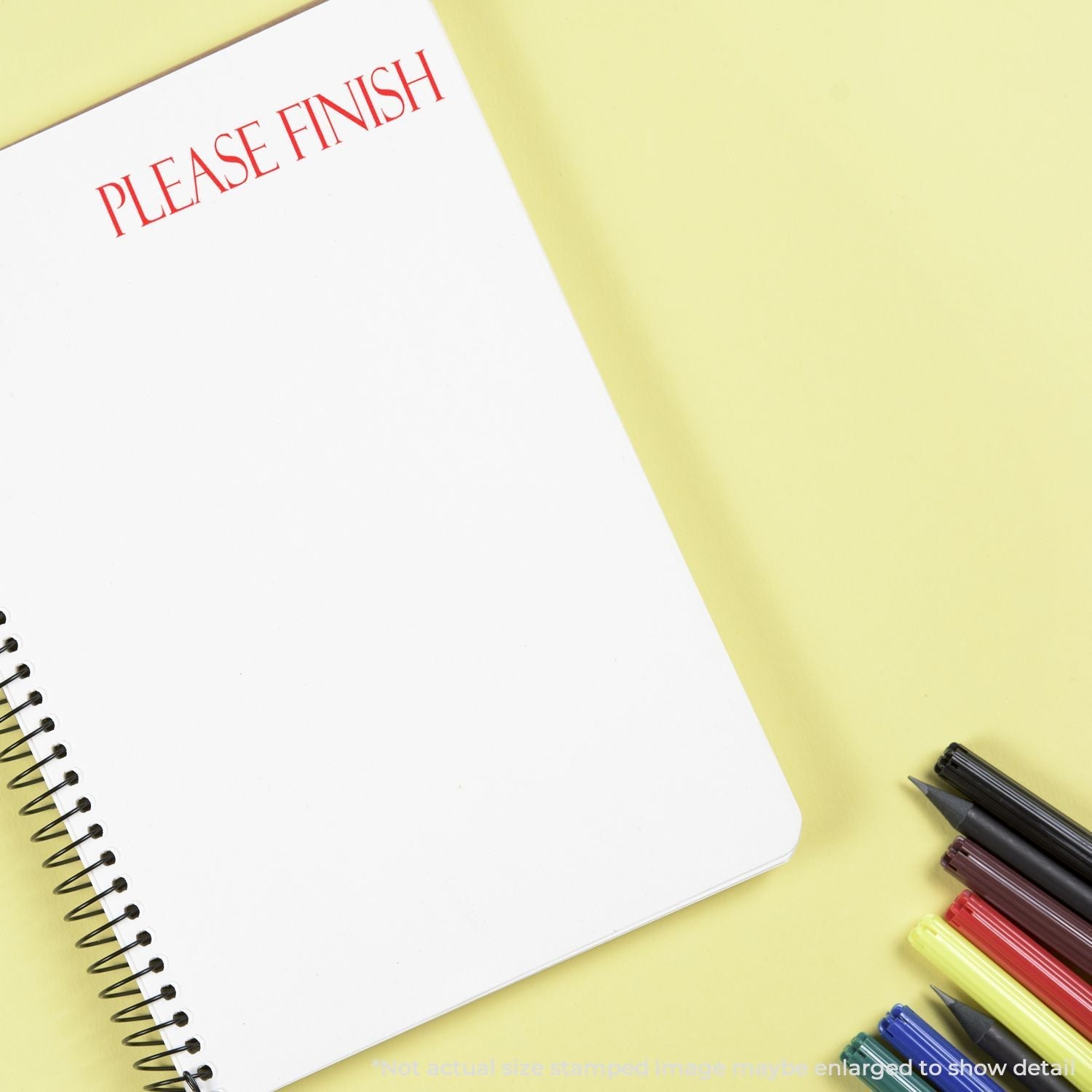 A notebook stamped with PLEASE FINISH using a Self Inking Please Finish Stamp, surrounded by colorful pens on a yellow background.