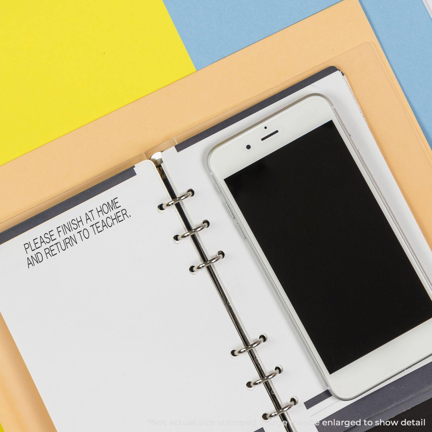 Please Finish At Home Return To Teacher Rubber Stamp on a notebook page next to a smartphone, with colorful background papers.