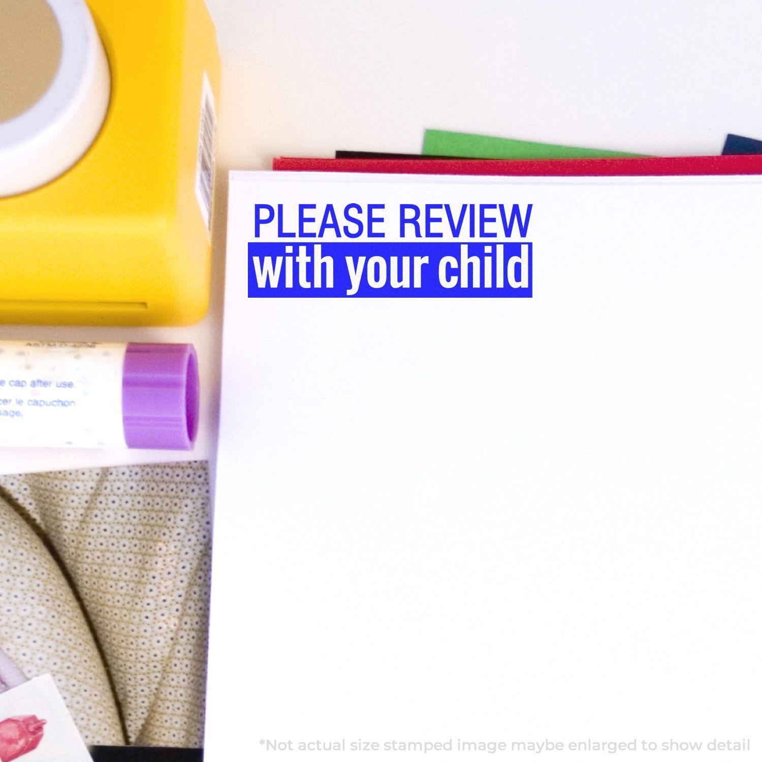 A stock office rubber stamp with a stamped image showing how the text "PLEASE REVIEW with your child" in a bold font and dual-colored marking is displayed after stamping.