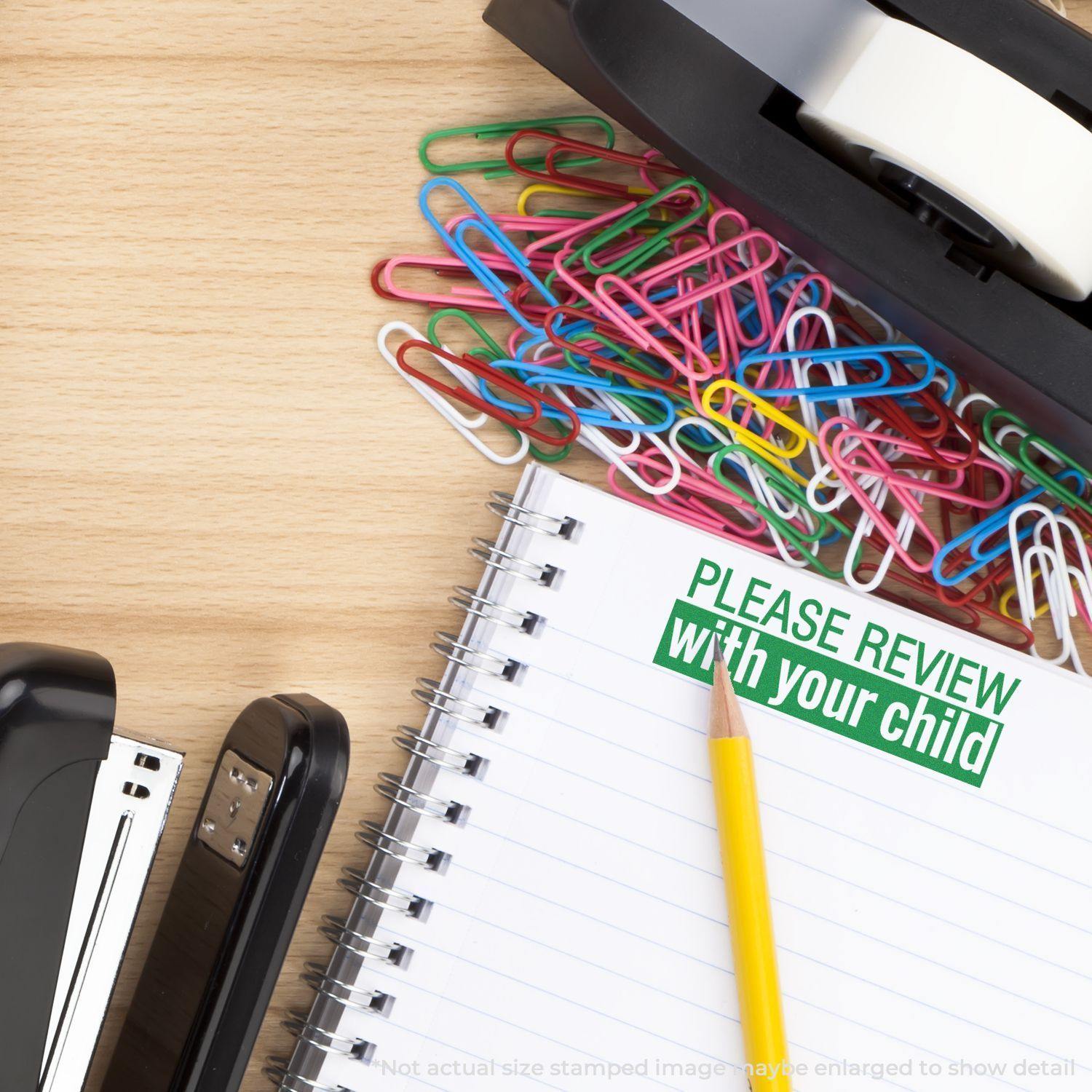 A desk with a stapler, tape dispenser, colorful paperclips, and a notebook stamped with Large Pre-Inked Please Review With Your Child Stamp in green.