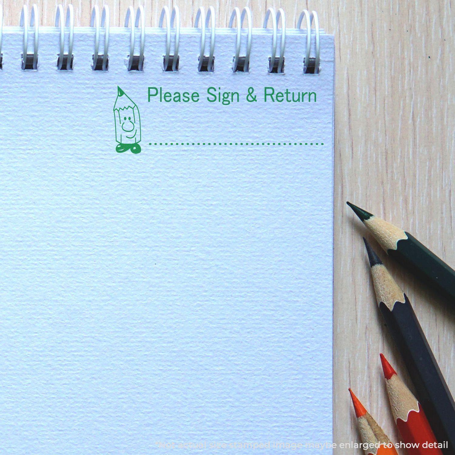 Large Self Inking Please Sign and Return Stamp in green ink on a spiral notebook, with colored pencils beside it.