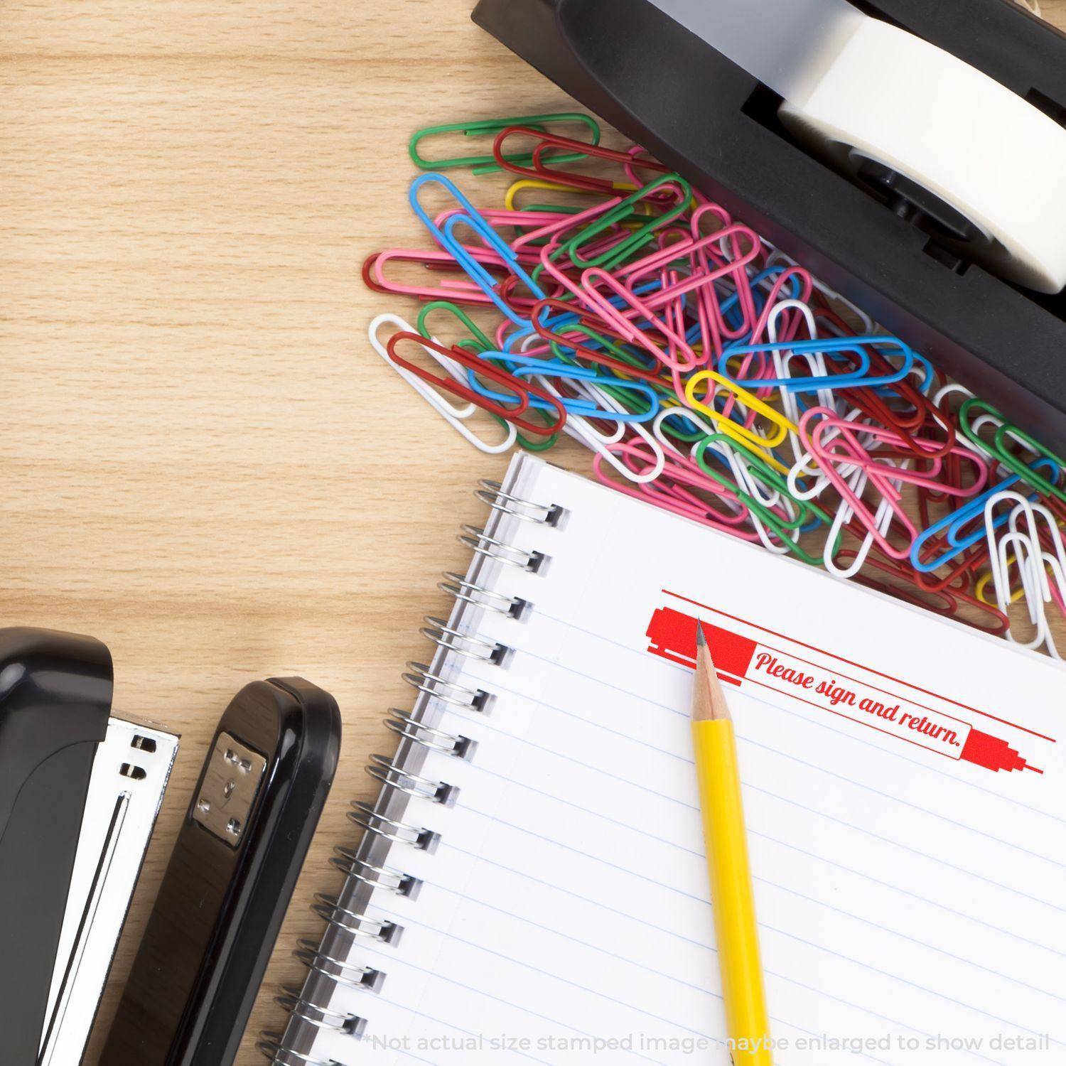 Large Please Sign and Return with Pen Pre-Inked Stamp used on a notebook page, surrounded by colorful paper clips, stapler, and tape dispenser.