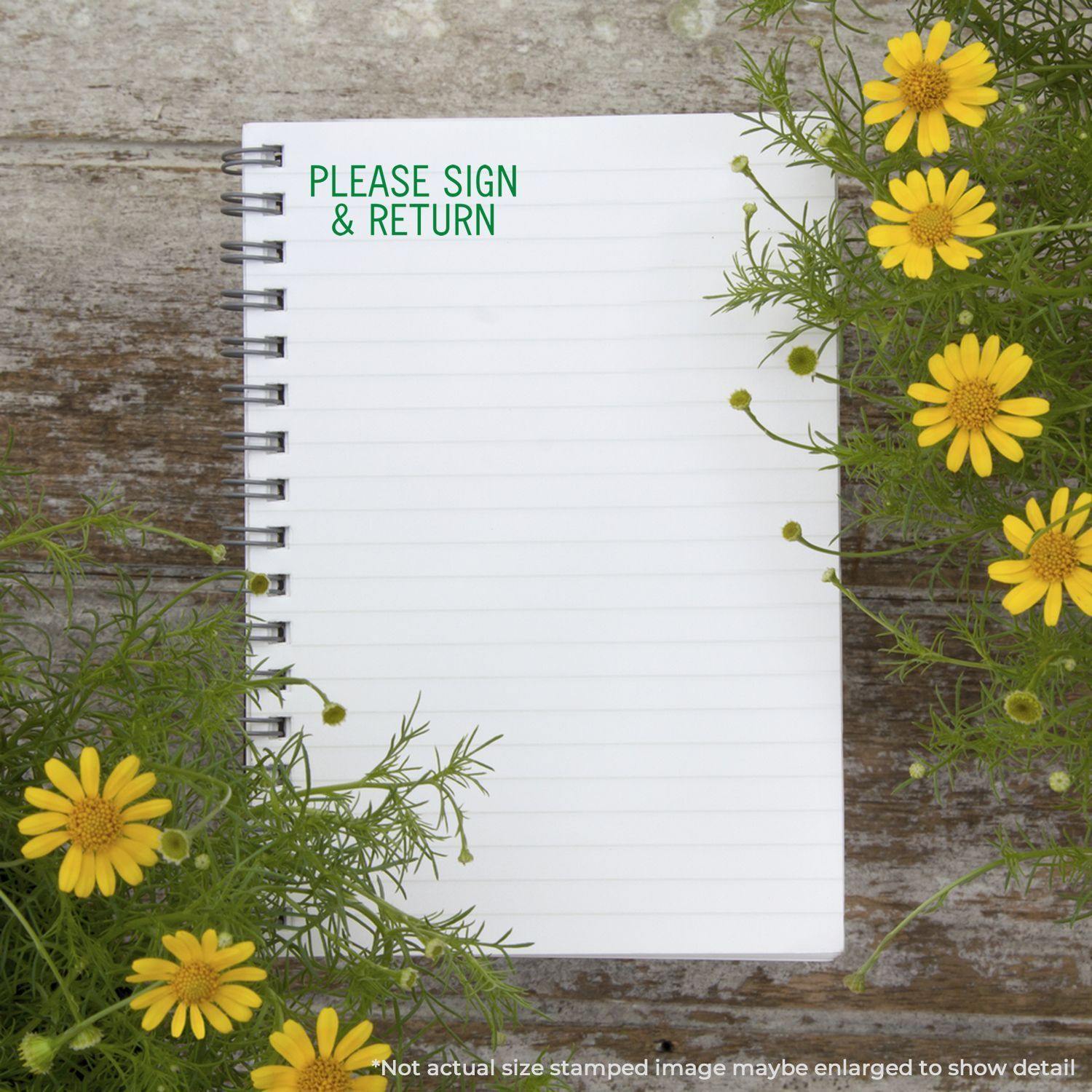 A notebook with a Please Sign & Return rubber stamp on a wooden surface, surrounded by yellow flowers and green foliage.