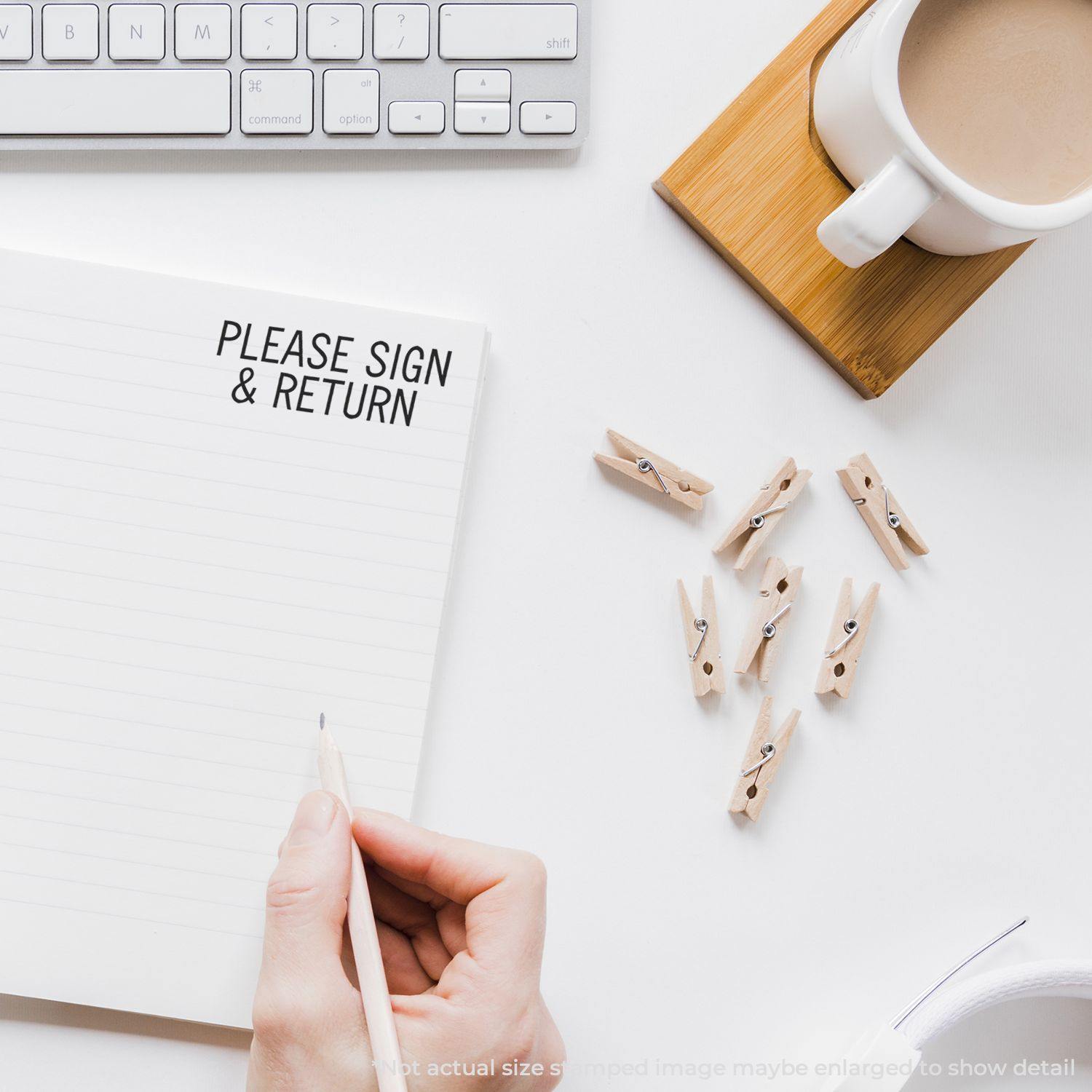 A hand holding a pen next to a notebook stamped with Please Sign & Return, with a keyboard, coffee cup, and clothespins nearby.