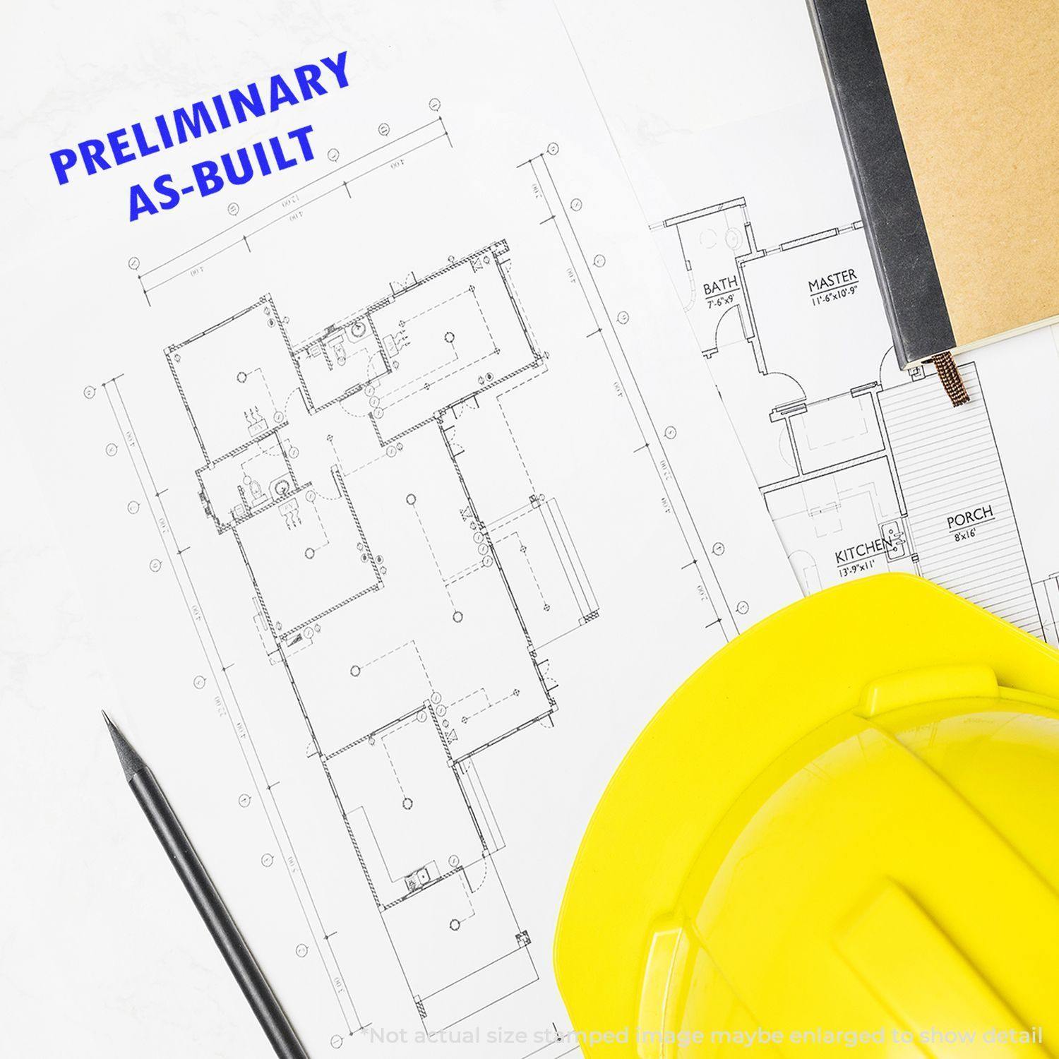 Preliminary As Built rubber stamp on architectural blueprint with a yellow hard hat, black pen, and notebook nearby.