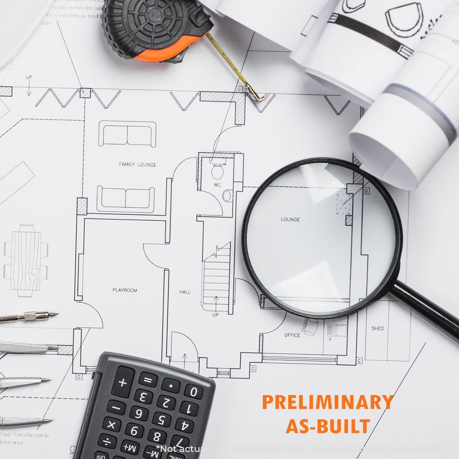 A Large Self Inking Preliminary As-Built Stamp is shown on a desk with architectural plans, a magnifying glass, a calculator, and a tape measure.