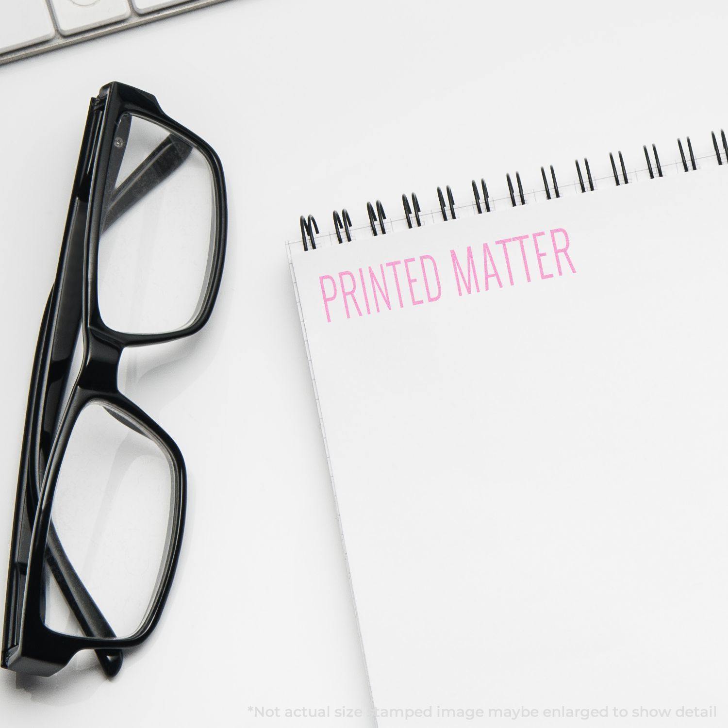 Large Pre-Inked Printed Matter Stamp used on a spiral notebook next to black glasses on a white desk.