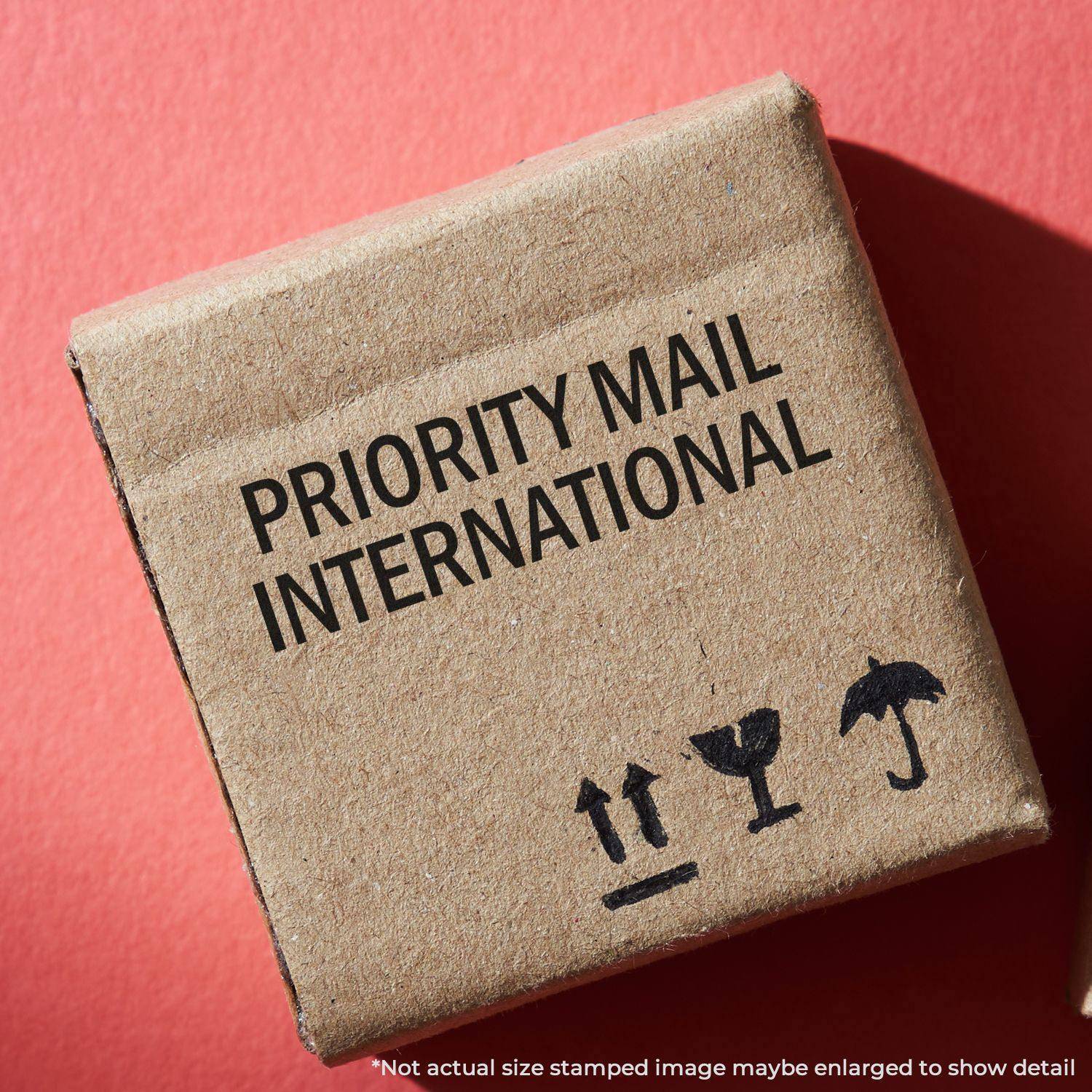 A cardboard box stamped with PRIORITY MAIL INTERNATIONAL using a Large Pre-Inked Priority Mail International Stamp on a red background.