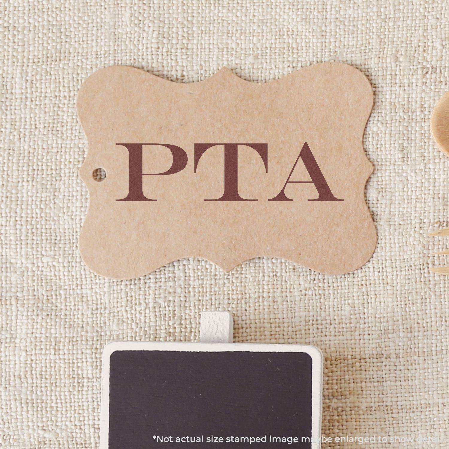 Large Pre-Inked PTA Stamp used on a brown tag with PTA text in bold letters, placed on a textured beige surface.