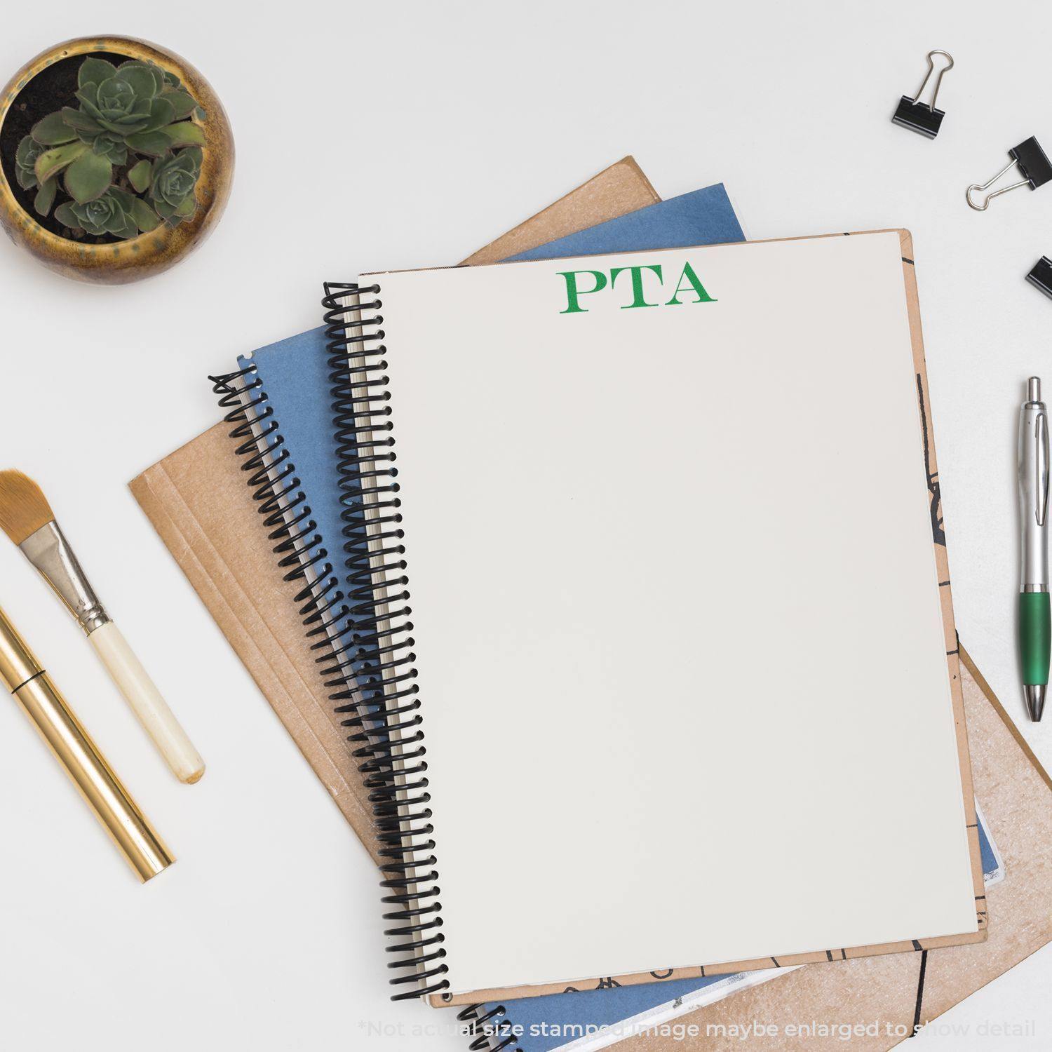 Large Pre-Inked PTA Stamp used on a spiral notebook, surrounded by office supplies including a pen, brushes, clips, and a plant.