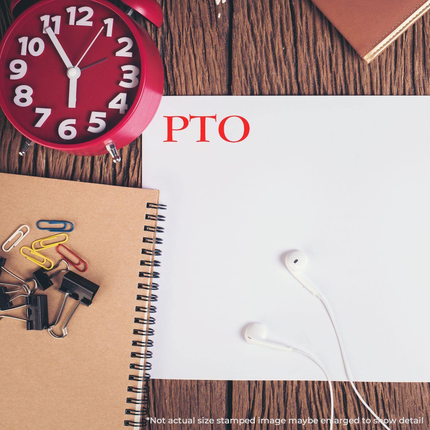 Large Pre-Inked PTO Stamp used on a white paper, surrounded by a red clock, notebooks, paper clips, and earphones on a wooden desk.