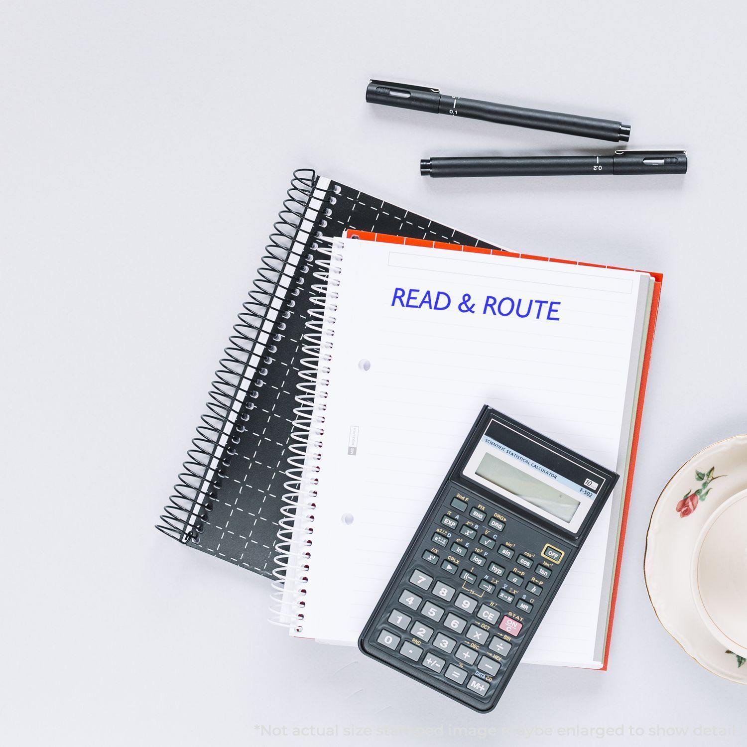 Large Pre-Inked Read & Route Stamp used on a notebook page, with pens, a calculator, and a teacup nearby on a white surface.