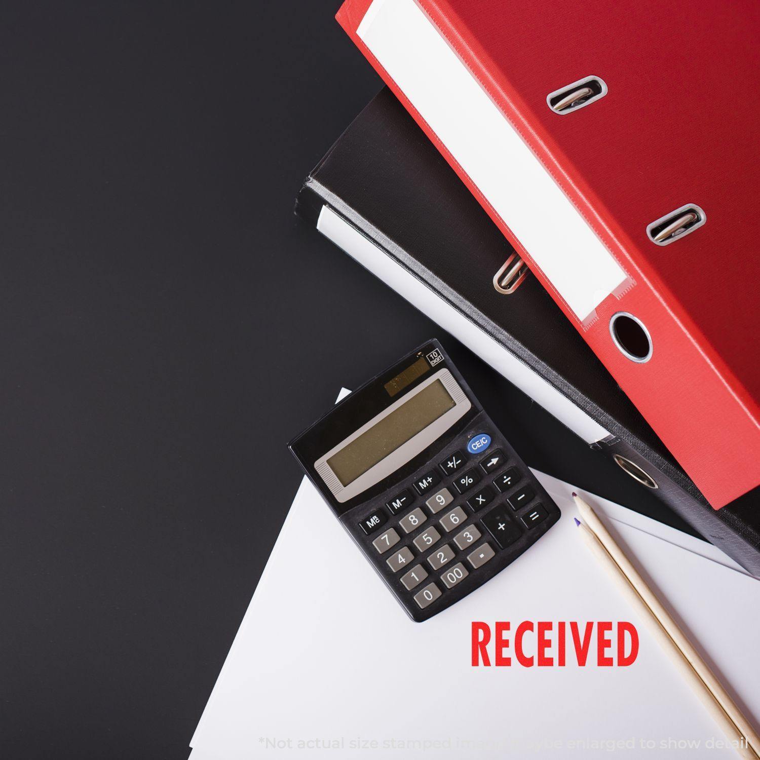 Large Received Rubber Stamp marking RECEIVED in red on paper, next to a calculator, pencil, and stacked binders on a black surface.