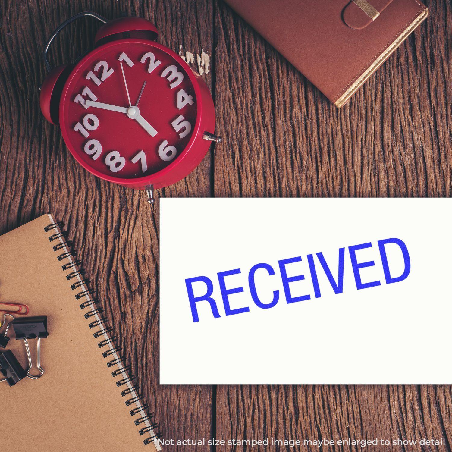 Large Pre-Inked Received Stamp used on a white paper, placed on a wooden desk with a red clock, notebook, and office supplies.