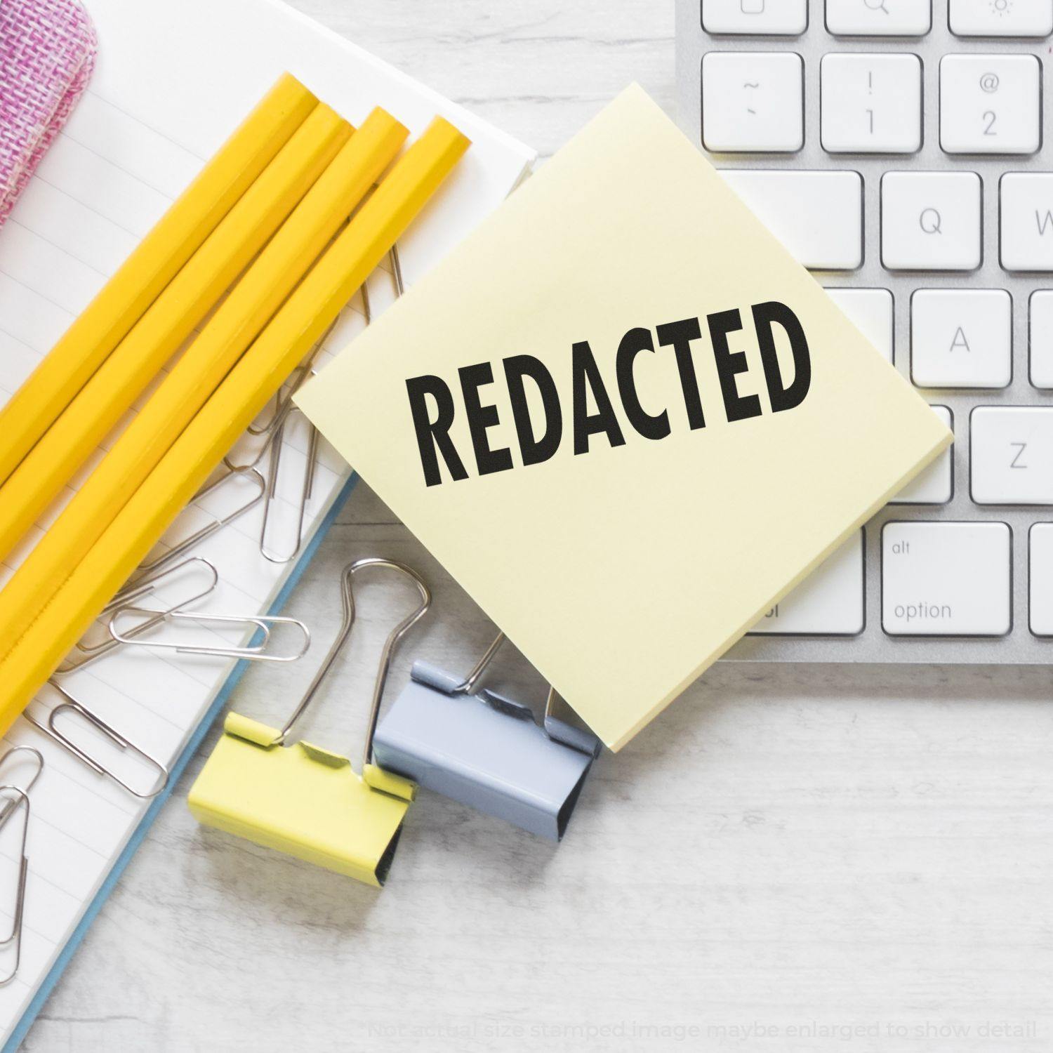 Large Redacted Rubber Stamp on a yellow sticky note, surrounded by pencils, paper clips, binder clips, and a keyboard on a desk.