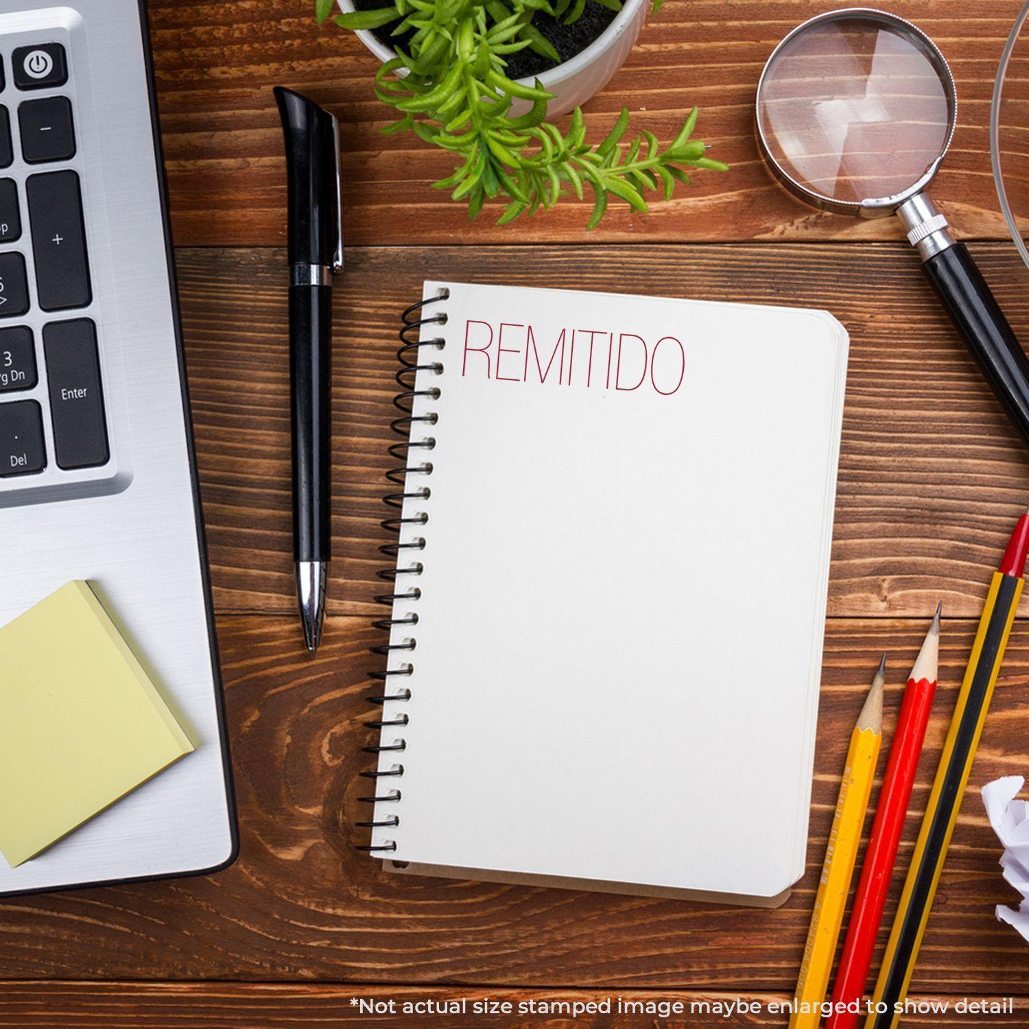 A Large Remitido Rubber Stamp is used on a spiral notebook placed on a wooden desk with a laptop, pen, sticky notes, and pencils.