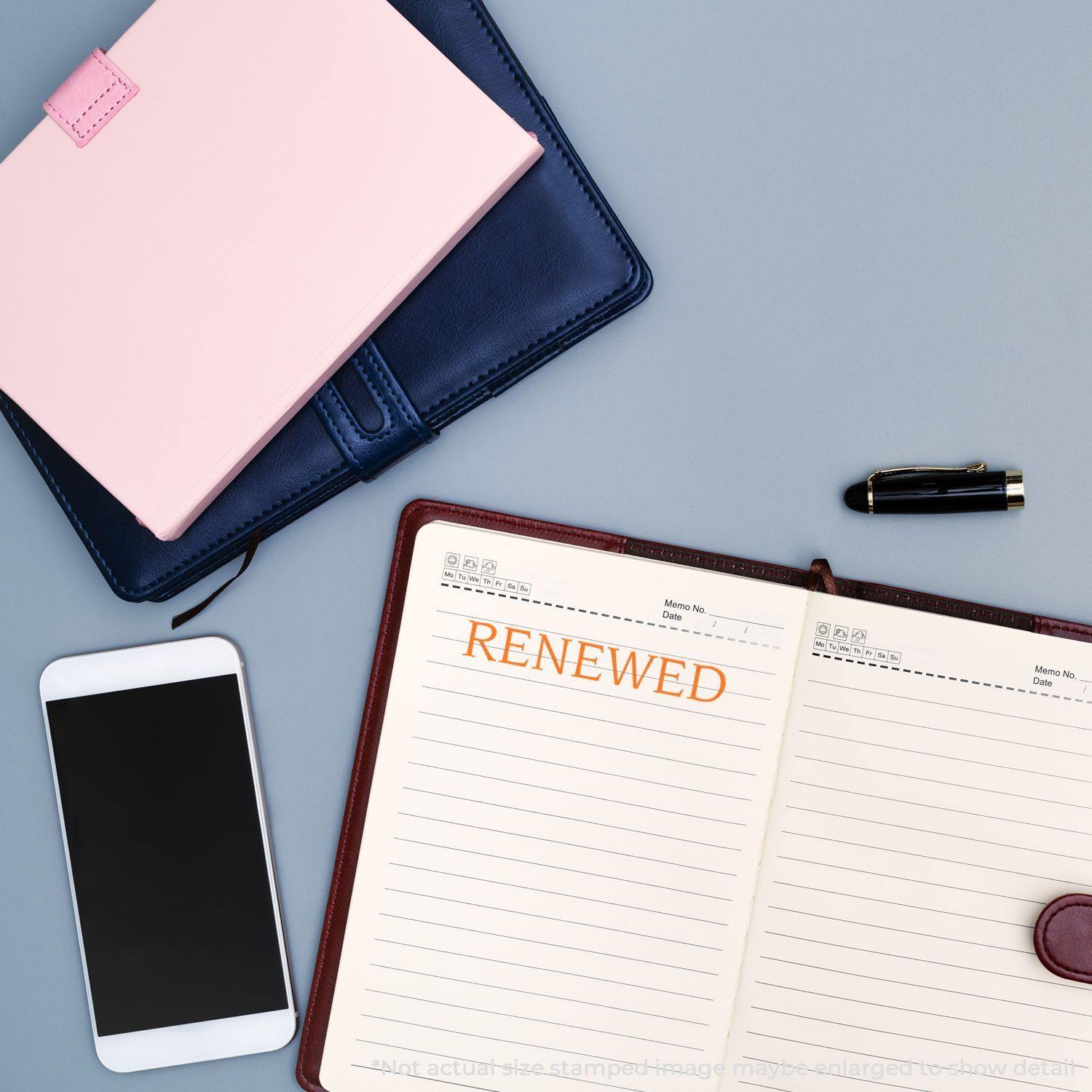 Large Pre-Inked Renewed Stamp used on an open planner, with a phone, pen, and closed notebooks nearby on a gray surface.