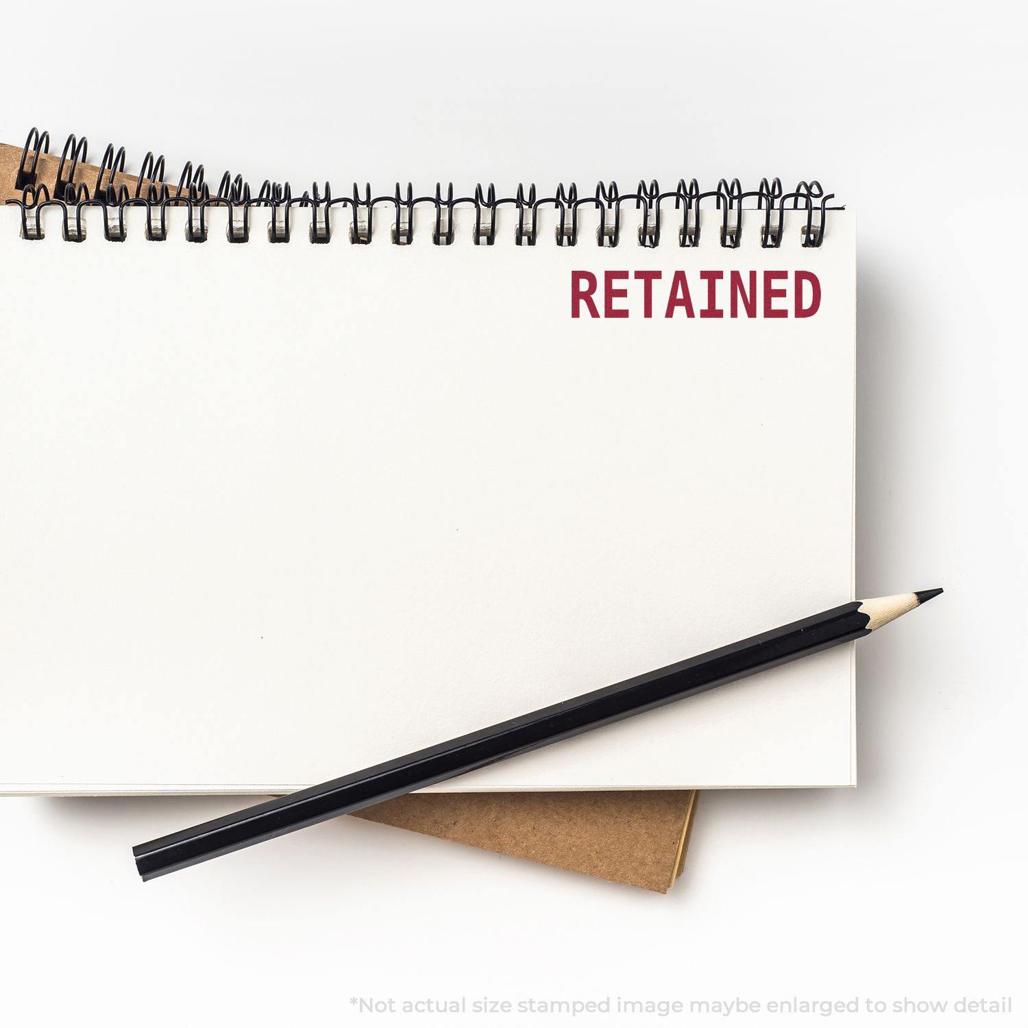 Large Self Inking Retained Stamp marking RETAINED in red on a white notebook page with a black pencil placed diagonally across the page.