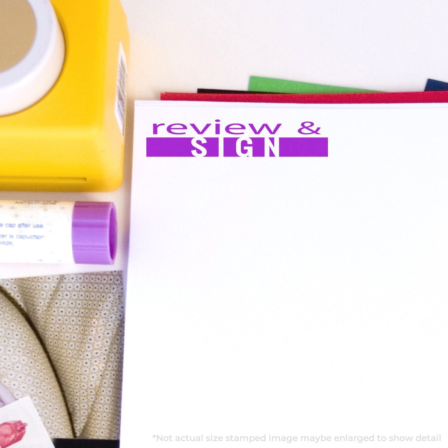 Self Inking Review and Sign Stamp in use on a white paper, surrounded by office supplies including a yellow stamp, glue stick, and folders.