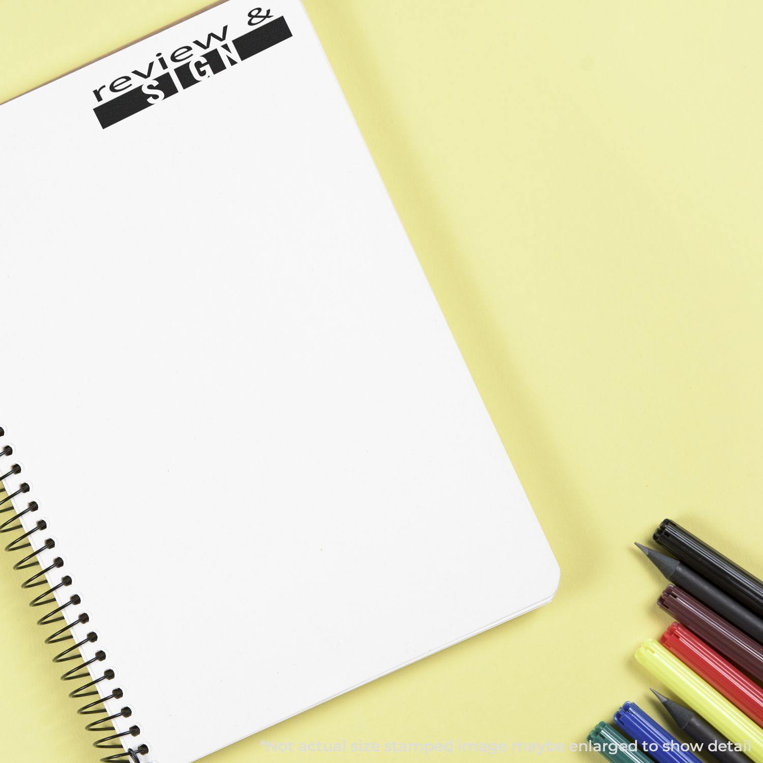 A Large Pre-Inked Review and Sign Stamp is used on a blank spiral notebook, with pens and markers placed beside it on a yellow surface.