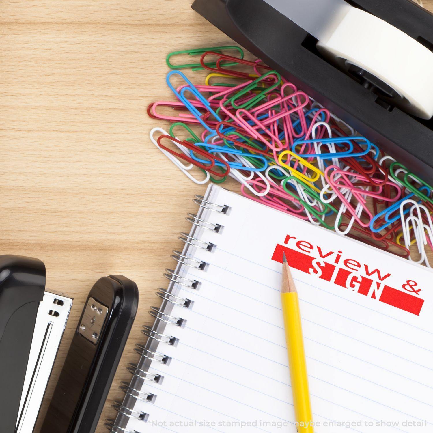 Large Review and Sign Rubber Stamp used on a notebook page with a pencil, stapler, tape dispenser, and colorful paper clips on a wooden desk.