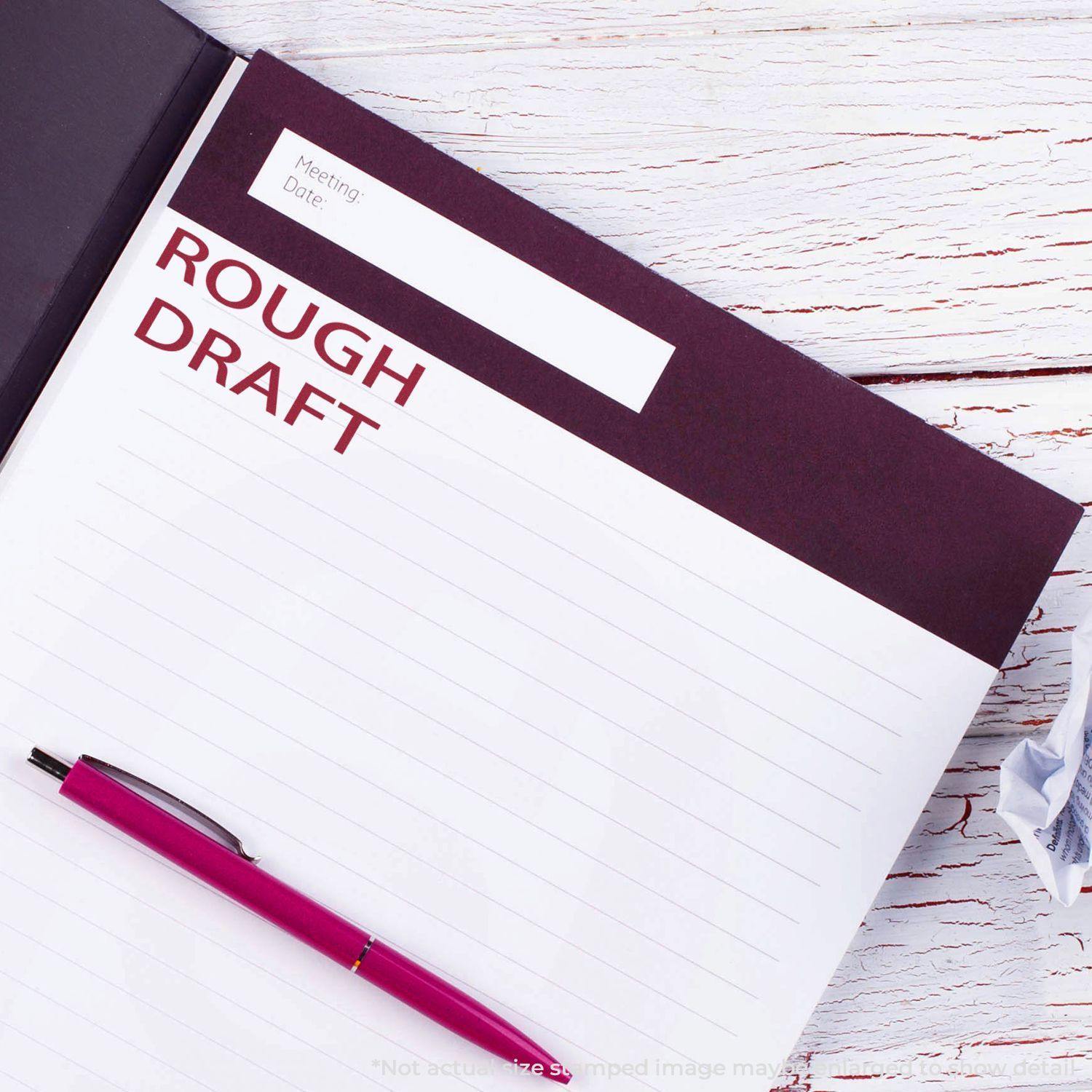 Large Self Inking Rough Draft Stamp used on a notebook page with ROUGH DRAFT stamped in red, next to a pink pen on a white wooden table.