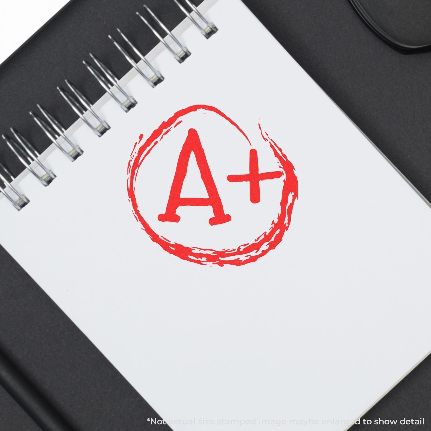 Self Inking Round A+ Stamp in red ink on a white spiral notebook, placed on a black surface.