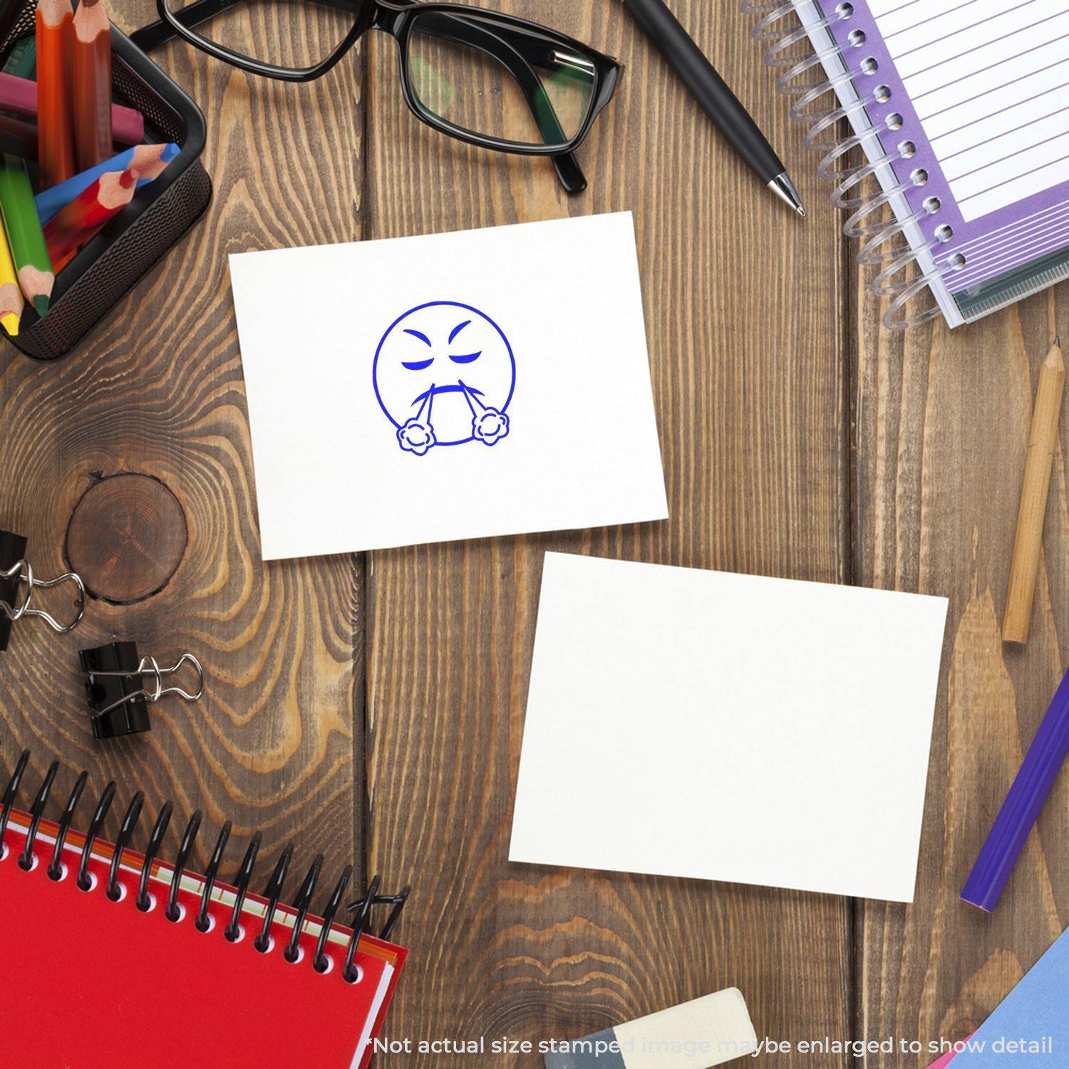 Round Angry Smiley Rubber Stamp on a white card, surrounded by office supplies like notebooks, glasses, and pens on a wooden desk.