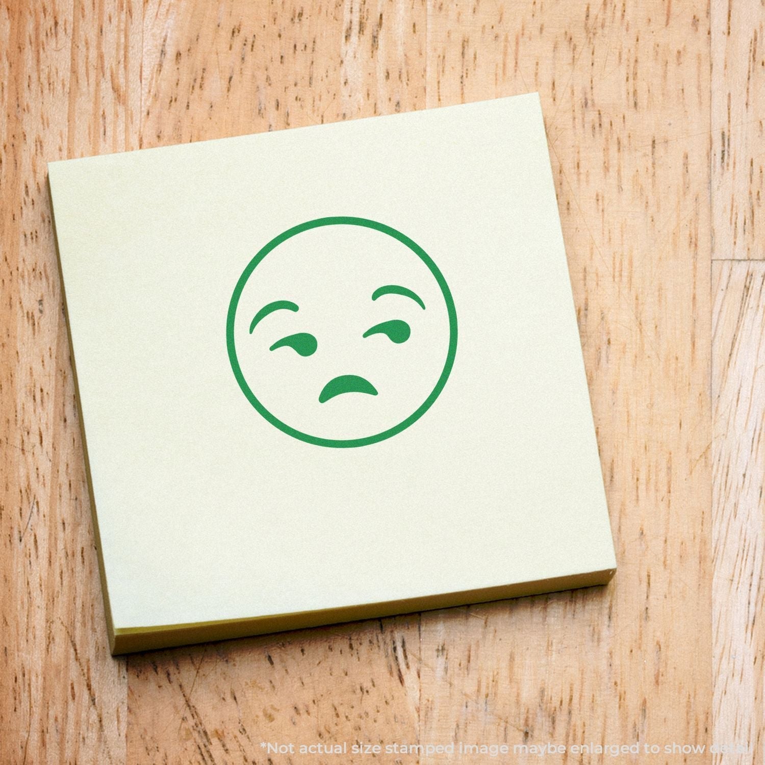 Self Inking Round Calm Smiley Stamp on a yellow sticky note, showing a calm, slightly disinterested face, placed on a wooden surface.