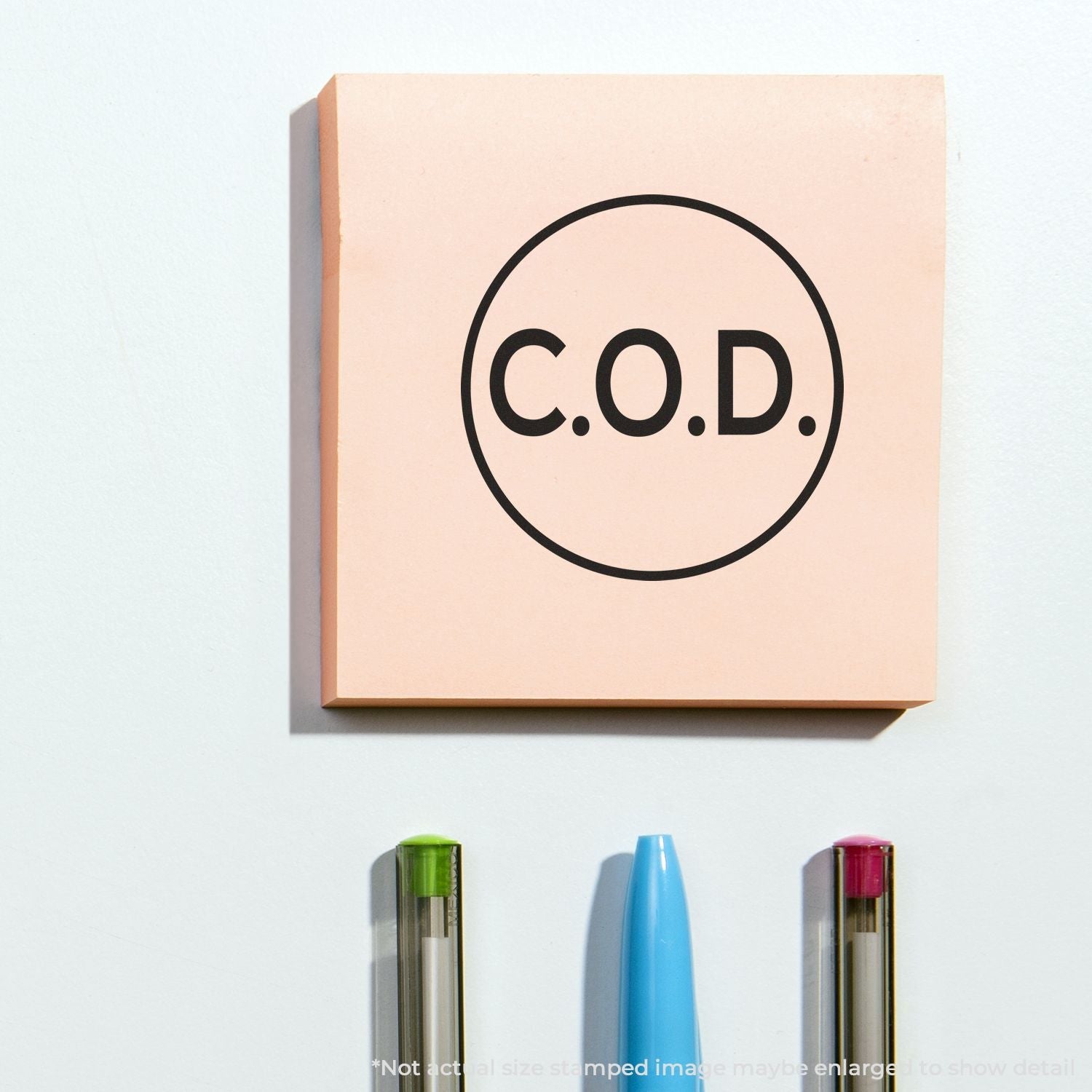 Self Inking Round COD Stamp used on a pink sticky note with "C.O.D." text, placed above three pens.