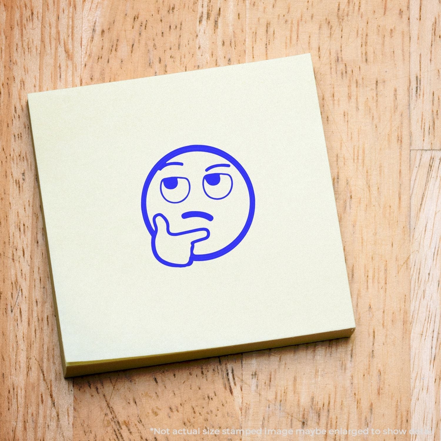 Self Inking Round Curious Smiley Stamp on a yellow sticky note, featuring a blue thinking face emoji, placed on a wooden surface.