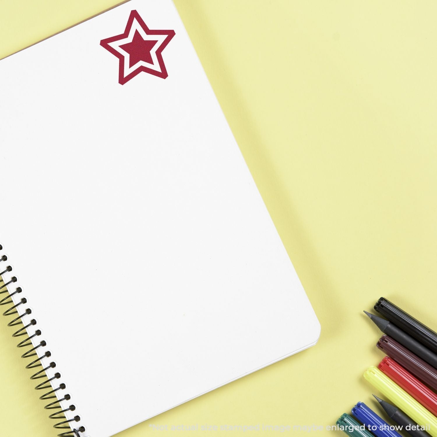 Self Inking Round Double Star Stamp in red ink on the top corner of an open spiral notebook, with colored markers beside it on a yellow surface.