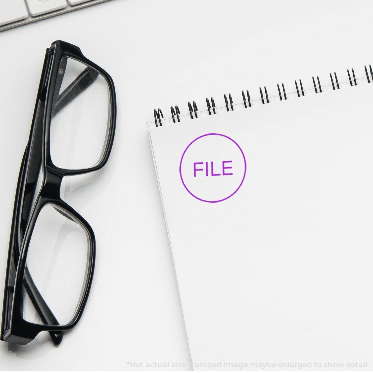 Round File Rubber Stamp on a spiral notebook with black glasses nearby on a white desk.