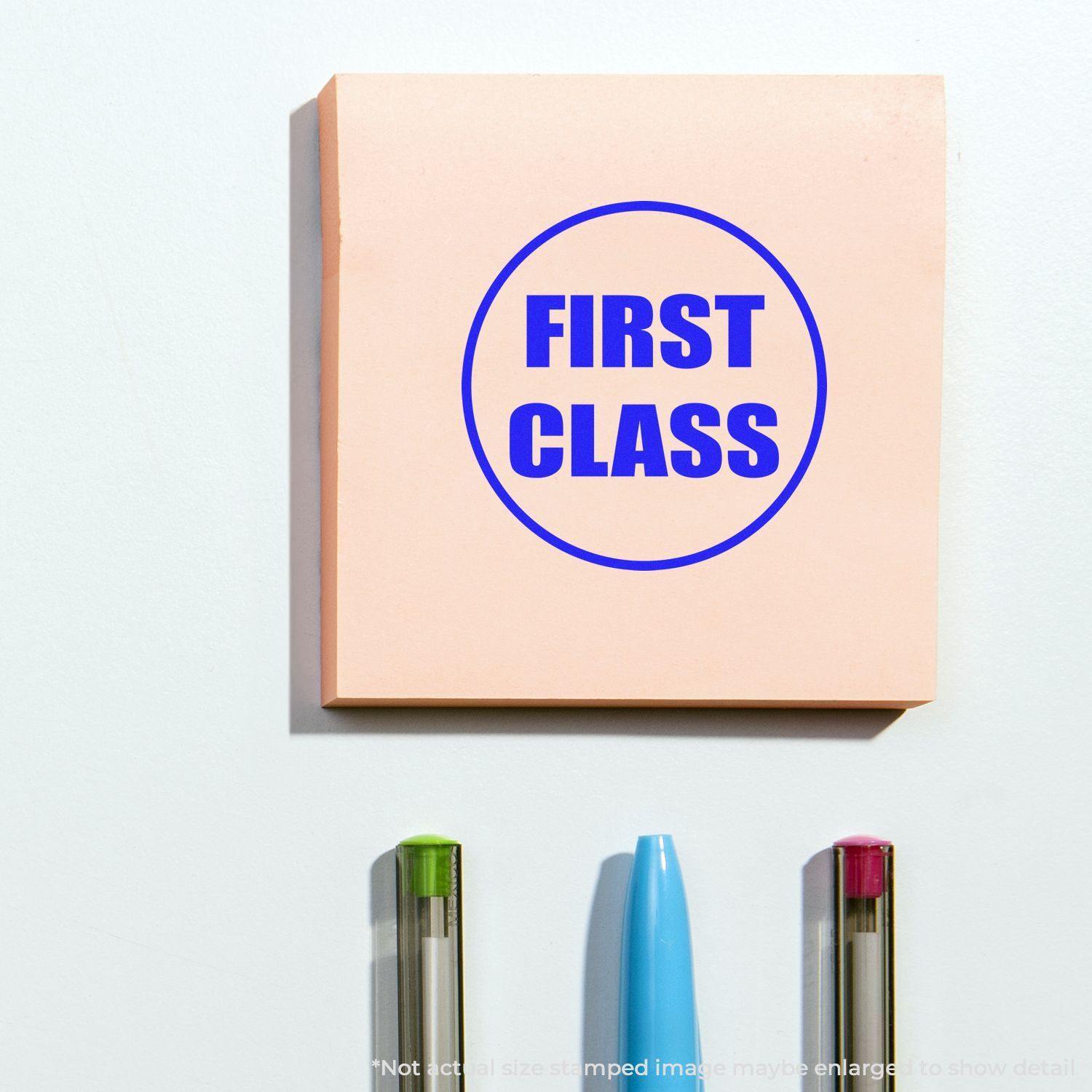 Round First Class rubber stamp impression on a pink sticky note, with three pens below it on a white surface.