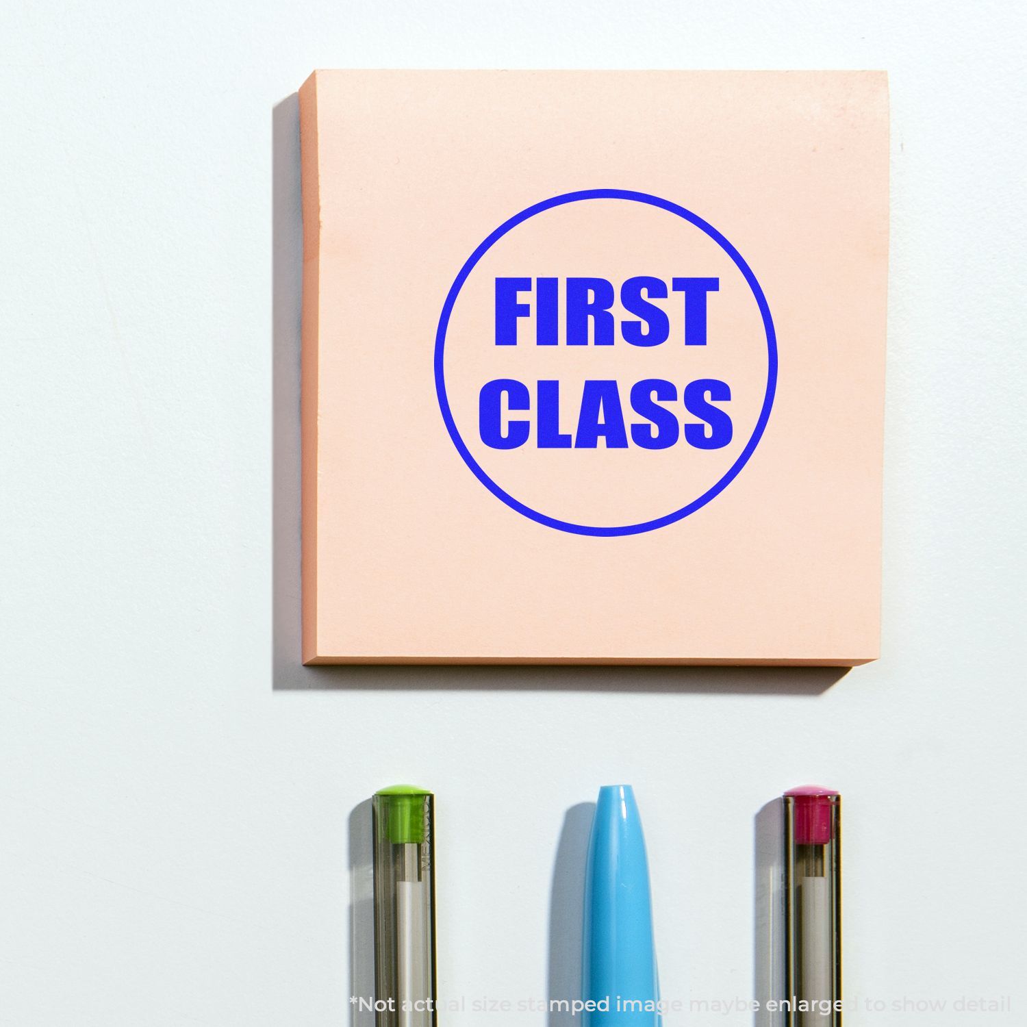 Self Inking Round First Class Stamp used on a pink sticky note with "FIRST CLASS" in blue, placed above three pens.