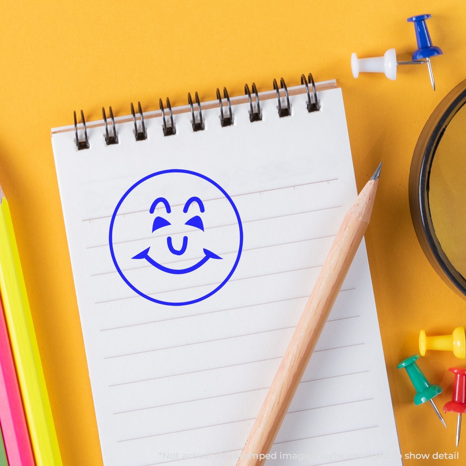 Self Inking Round Happy Face Stamp in blue ink on a notepad, surrounded by a pencil, colorful push pins, and stationery on a yellow background.