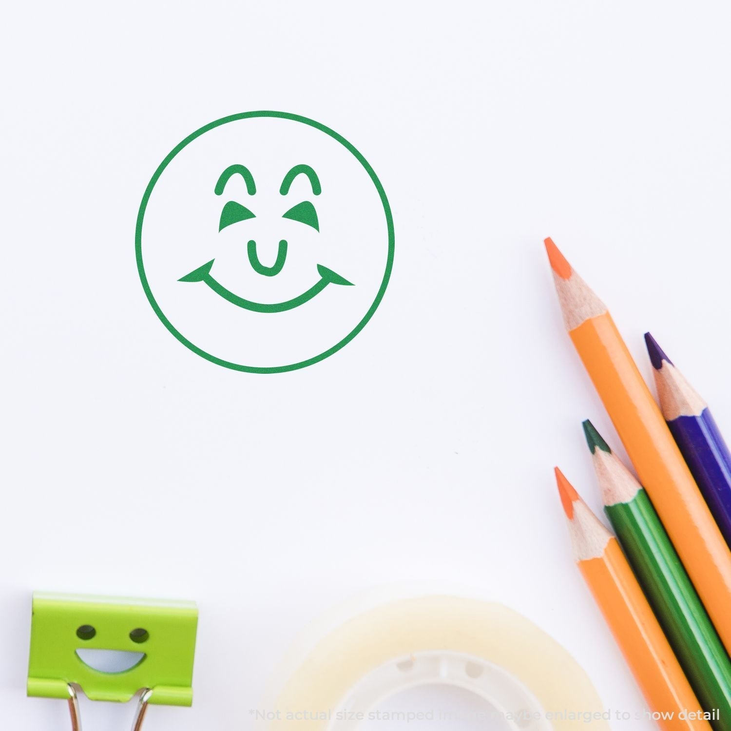 Self Inking Round Happy Face Stamp in green ink on white paper, with colored pencils, tape, and a smiley face paper clip nearby.