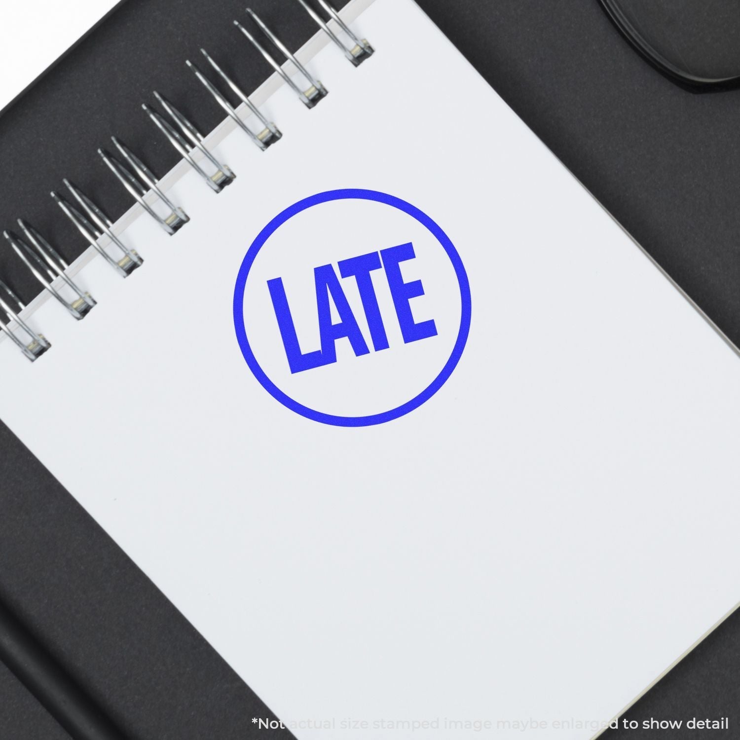 A Self Inking Round Late Stamp imprint in blue ink on a white spiral notebook, placed on a black surface.