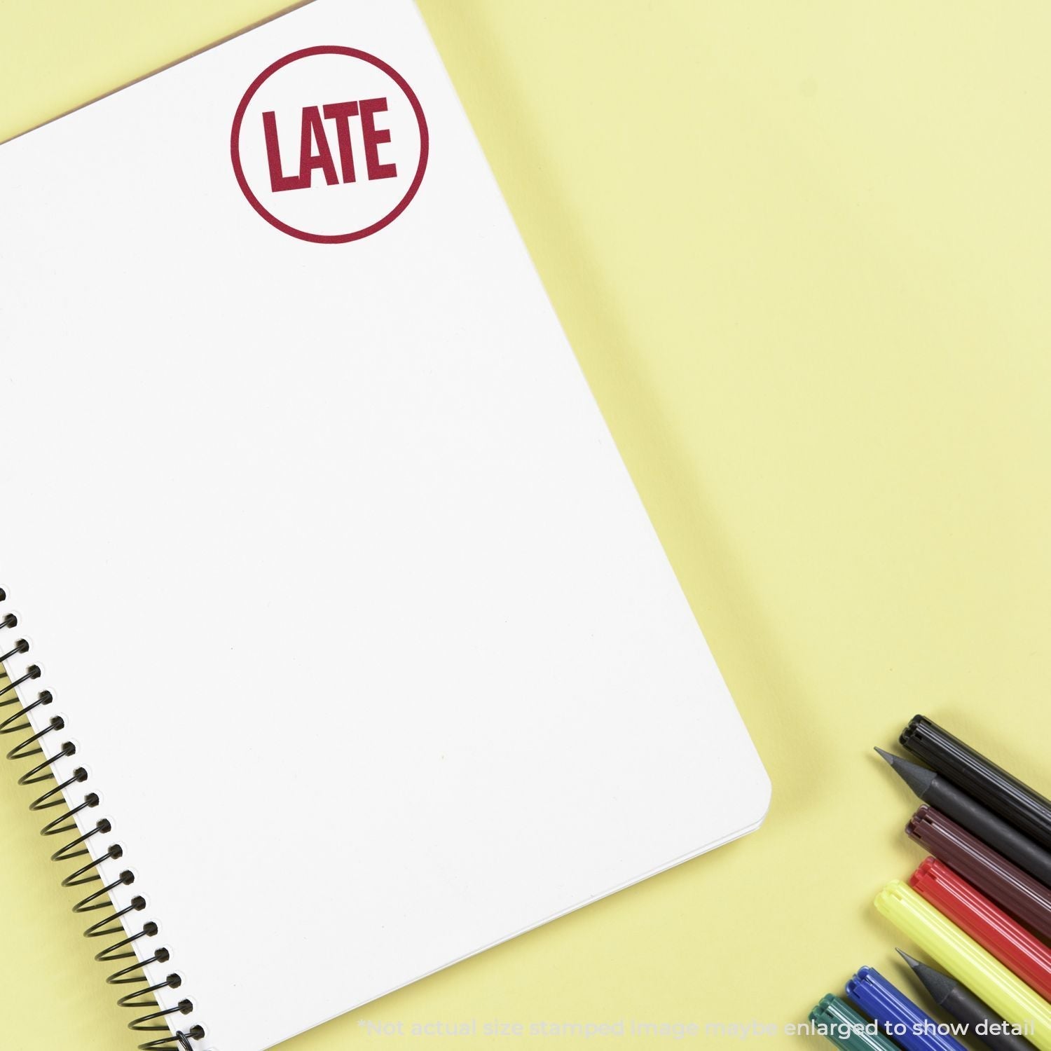 Self Inking Round Late Stamp marking LATE on a white spiral notebook, with pens and highlighters on a yellow background.