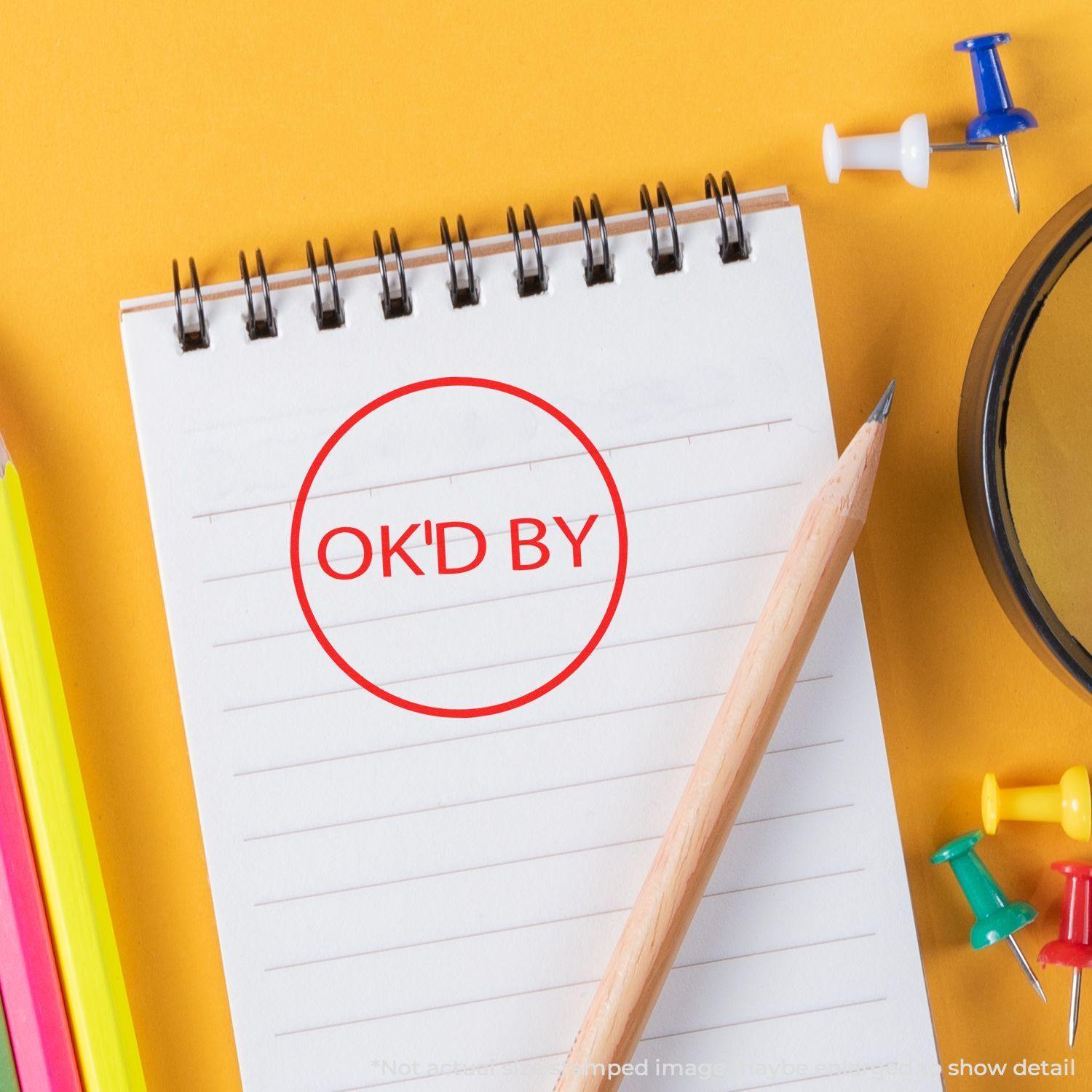 A notepad with a red Round Ok'd By Rubber Stamp imprint, surrounded by a pencil, push pins, and colorful stationery on a yellow background.