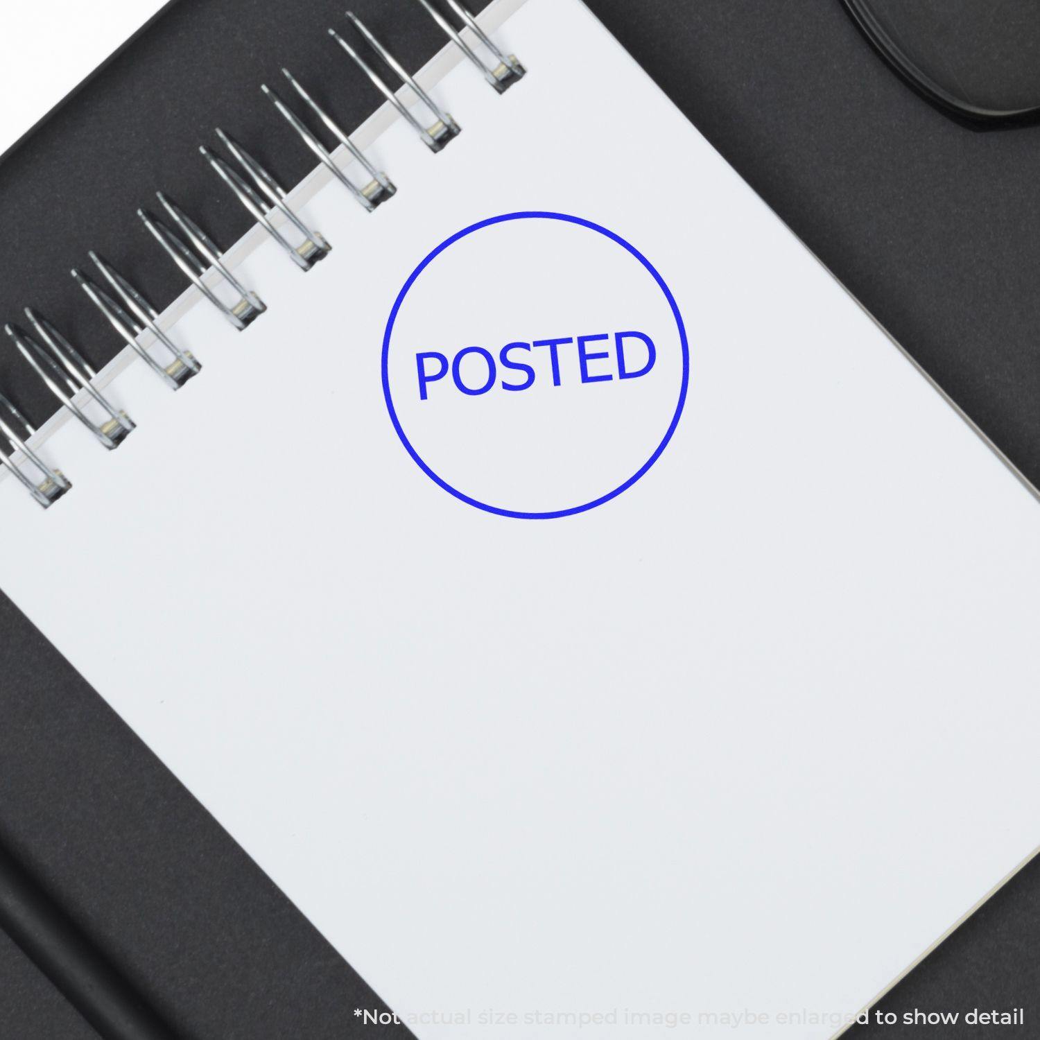 Round Posted Rubber Stamp marked in blue ink on a white spiral notebook page, placed on a black surface.