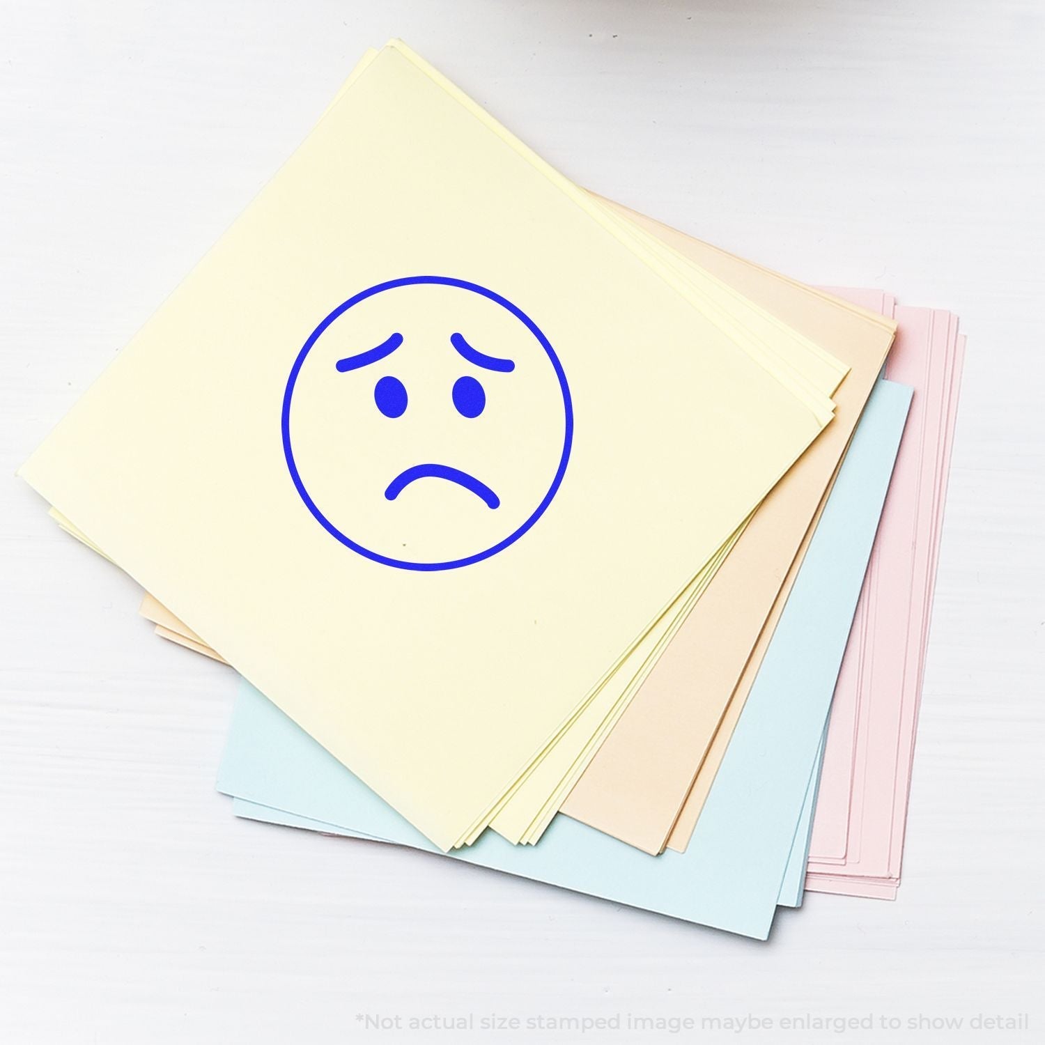 Self Inking Round Sad Face Stamp on a stack of pastel-colored papers, displaying a blue sad face impression.