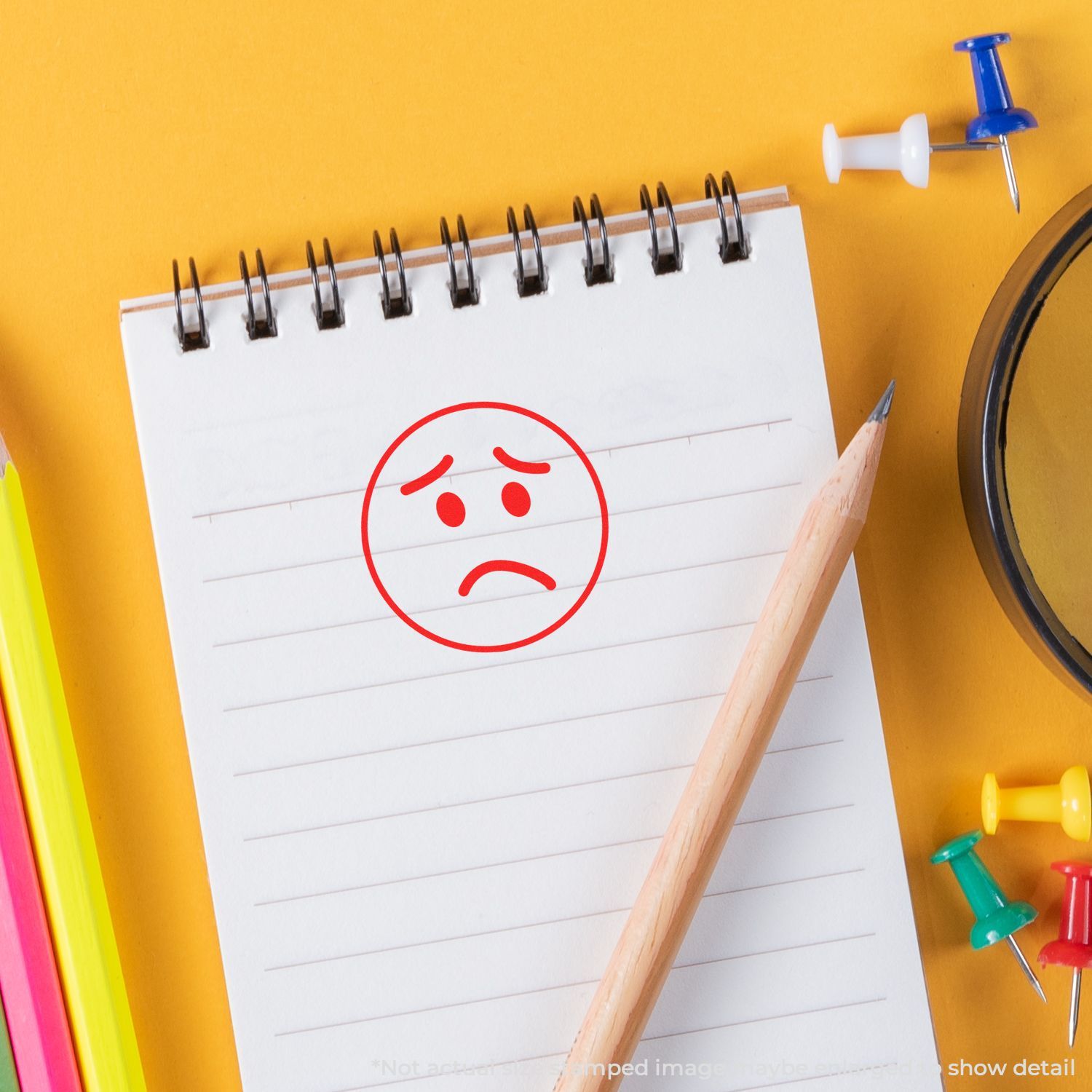 Self Inking Round Sad Face Stamp on a notepad with a pencil, colorful push pins, and a magnifying glass on a yellow background.