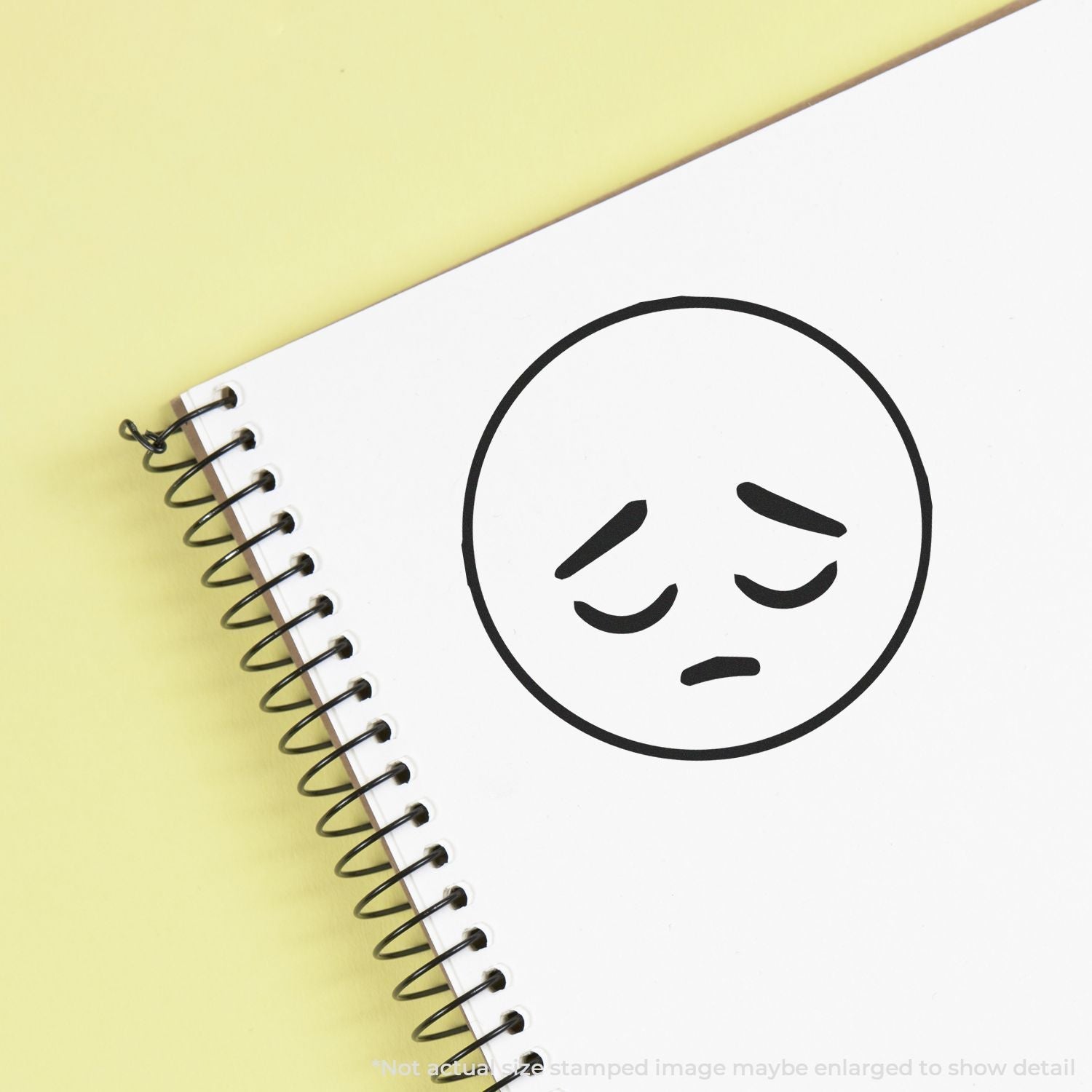 Self Inking Round Sad Smiley Stamp on a spiral notebook with a sad face impression on a yellow background.