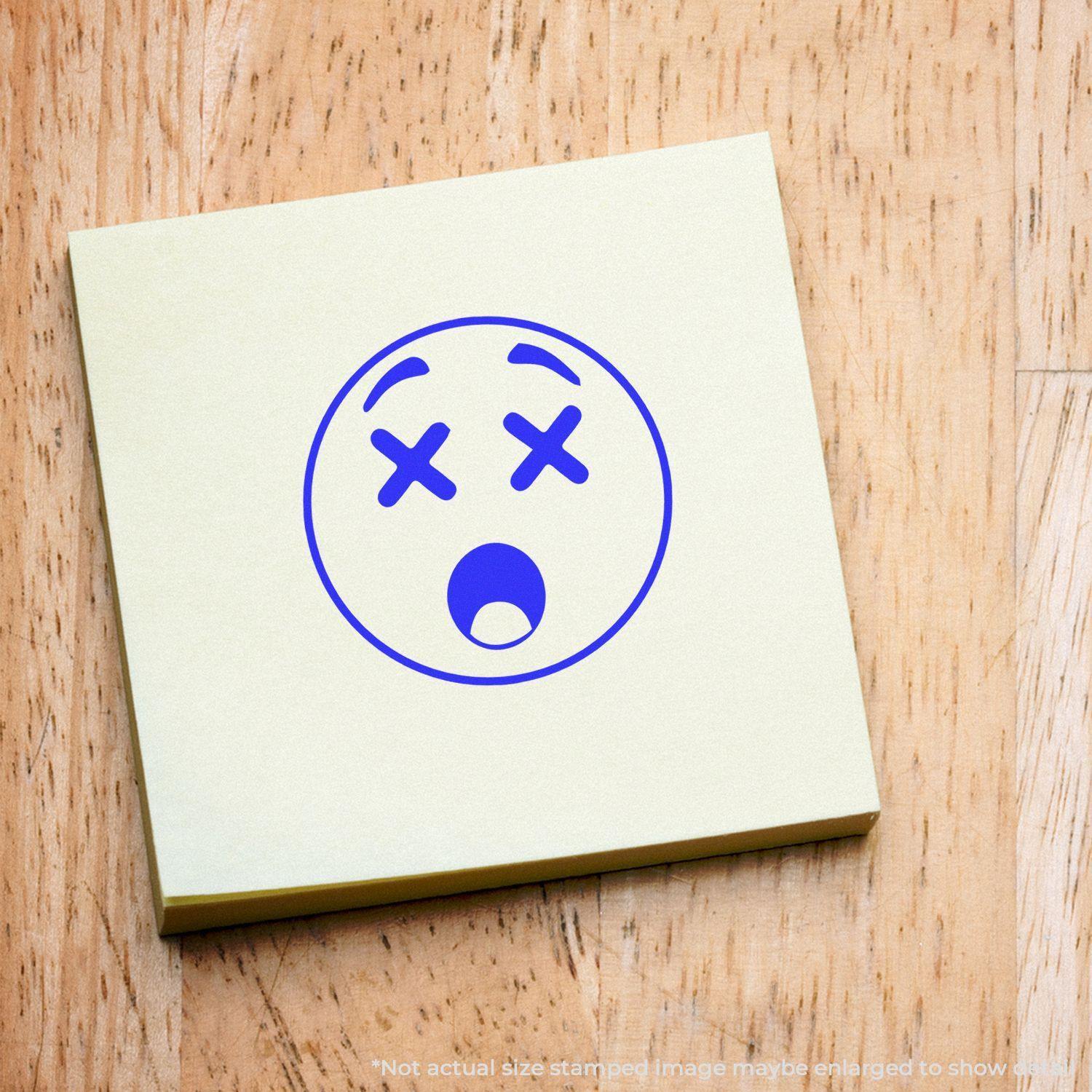 Round Shocked Smiley Rubber Stamp on a yellow sticky note with a wooden background, showing a surprised face with X eyes and open mouth.