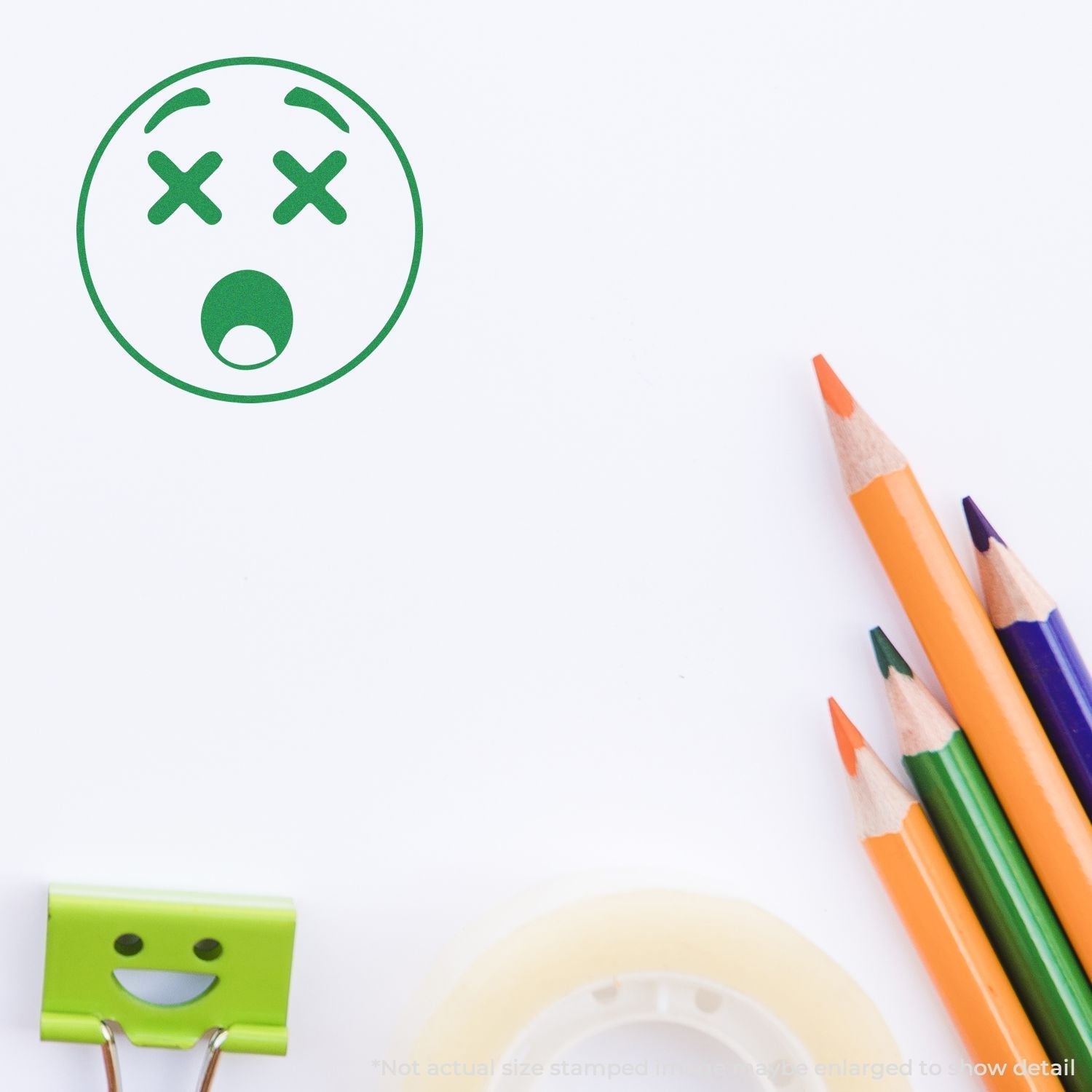 Self Inking Round Shocked Smiley Stamp impression next to colorful pencils, a green binder clip, and a roll of tape on a white surface.