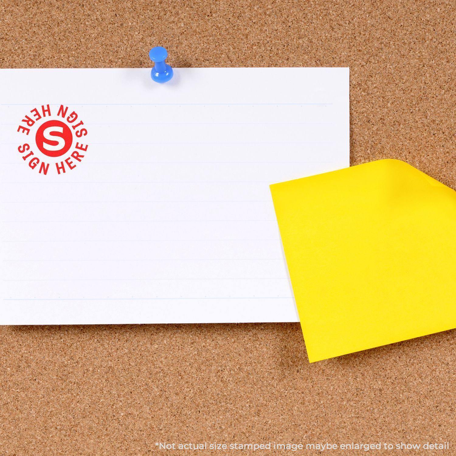 Round Sign Here Sign Here Rubber Stamp on a white paper pinned to a corkboard, with a yellow sticky note beside it.