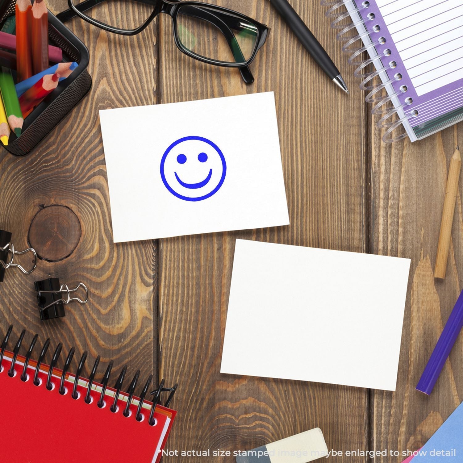 A Self Inking Round Smiley Stamp is used on a white card, surrounded by office supplies like notebooks, glasses, and pens on a wooden desk.