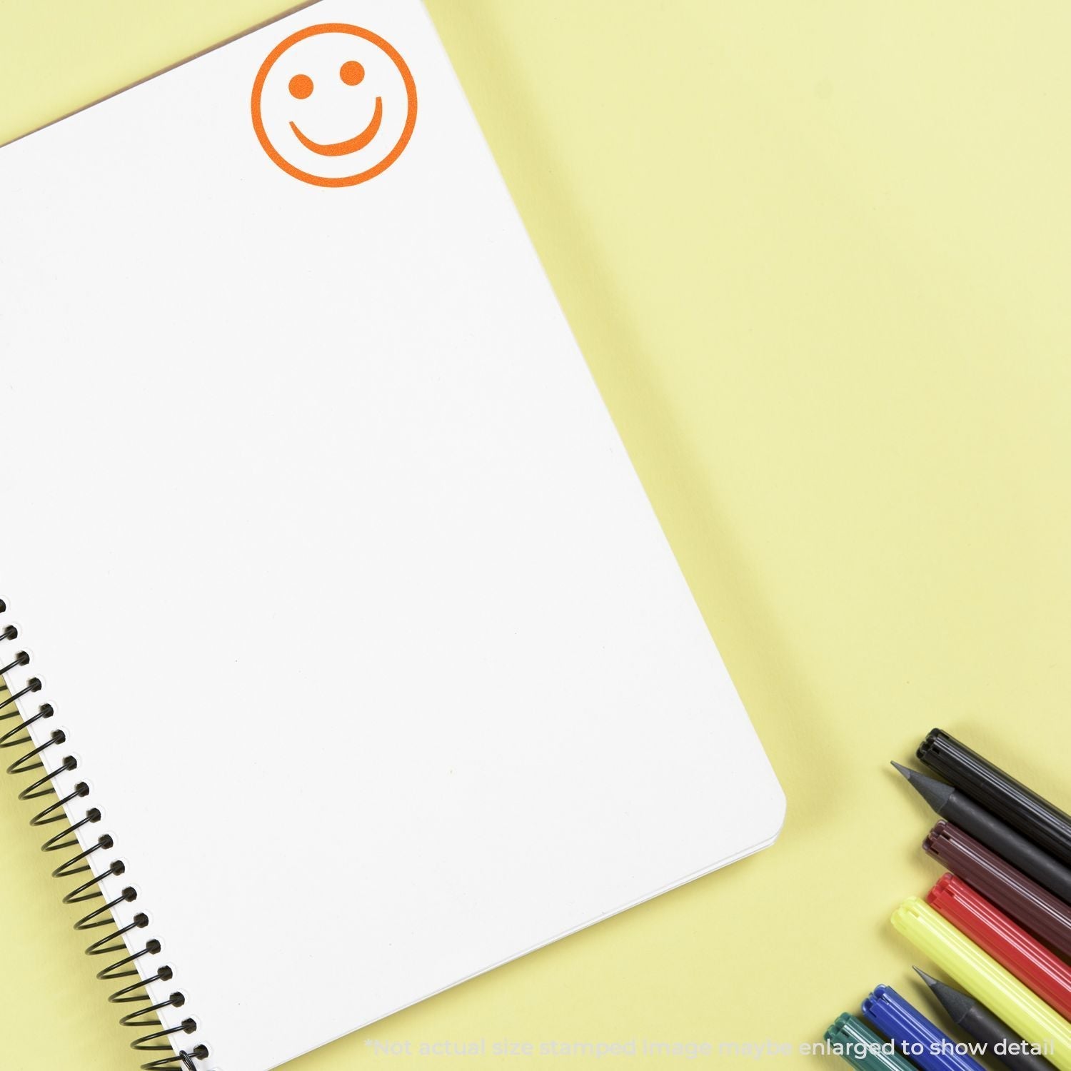 A Self Inking Round Smiley Stamp imprint on a blank spiral notebook page, with colorful pens nearby on a yellow background.