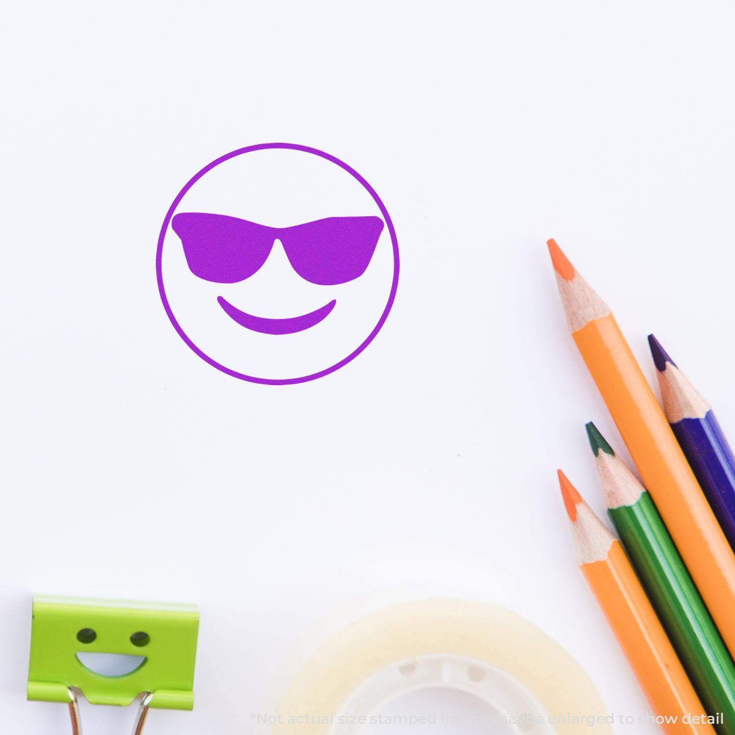 Round Smiley with Sunglasses Rubber Stamp in purple ink on white paper, surrounded by colorful pencils, a clip, and tape.